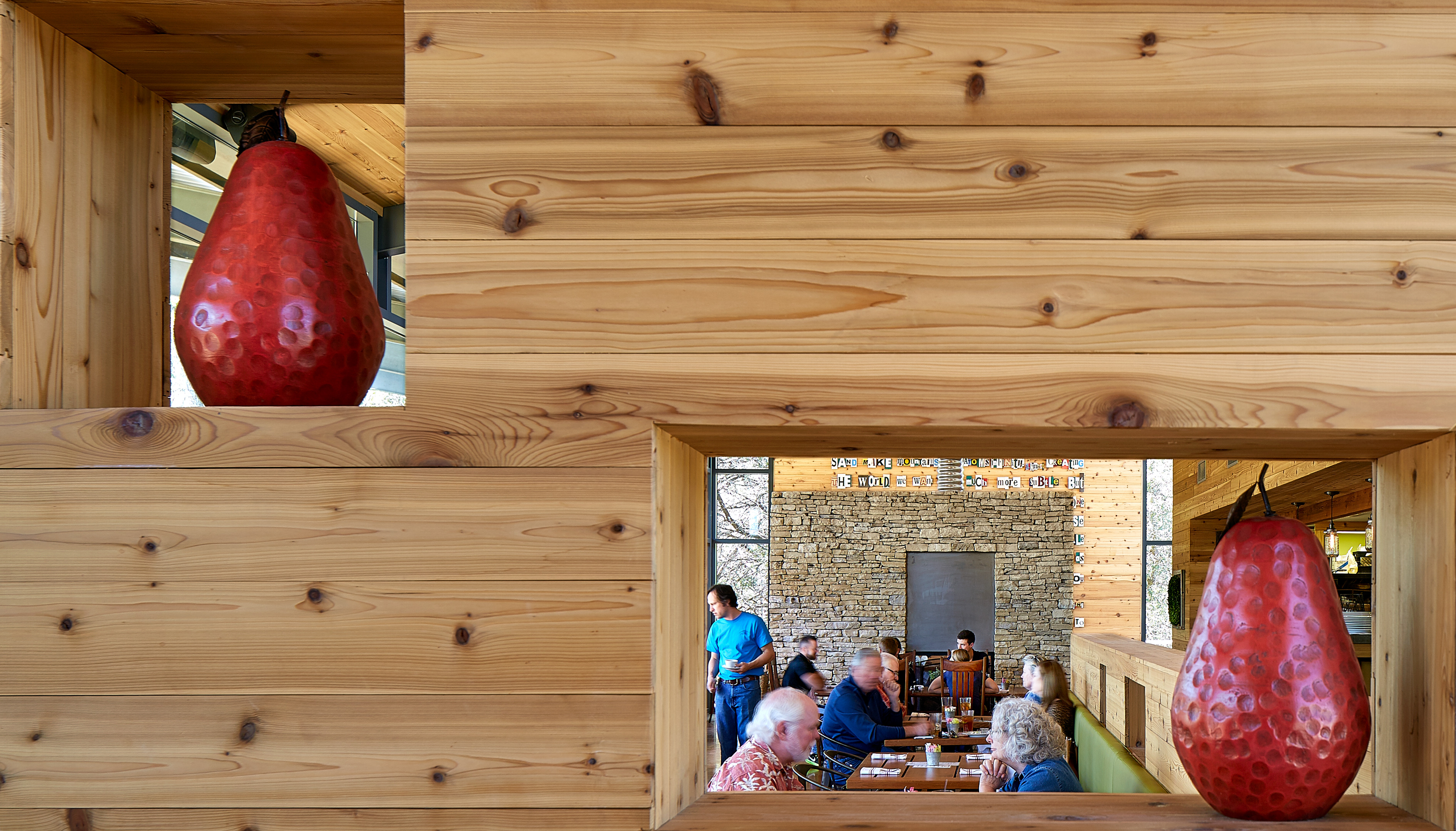 A wooden partition with cutouts displaying red pear-shaped sculptures separates diners sitting at tables in a restaurant. The background shows more patrons and part of a stone wall, creating an inviting atmosphere that feels casually orchestrated, almost as if by Auto Draft.