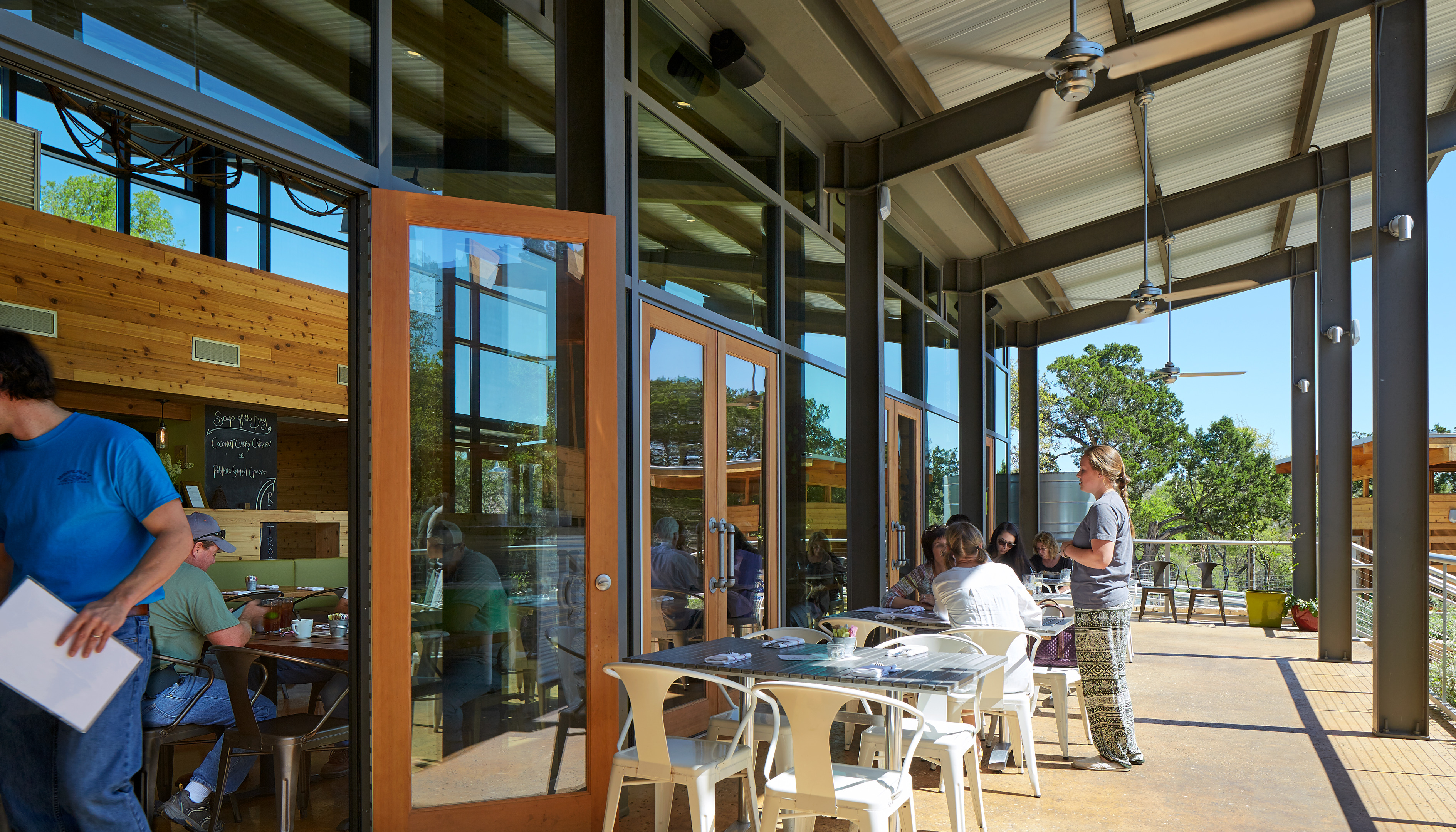 Outdoor dining area with several people sitting at tables under a covered patio. A waiter in a blue shirt is serving customers inside the adjacent restaurant, which has large glass doors dividing the spaces. The welcoming atmosphere feels seamless, as if created by auto draft.