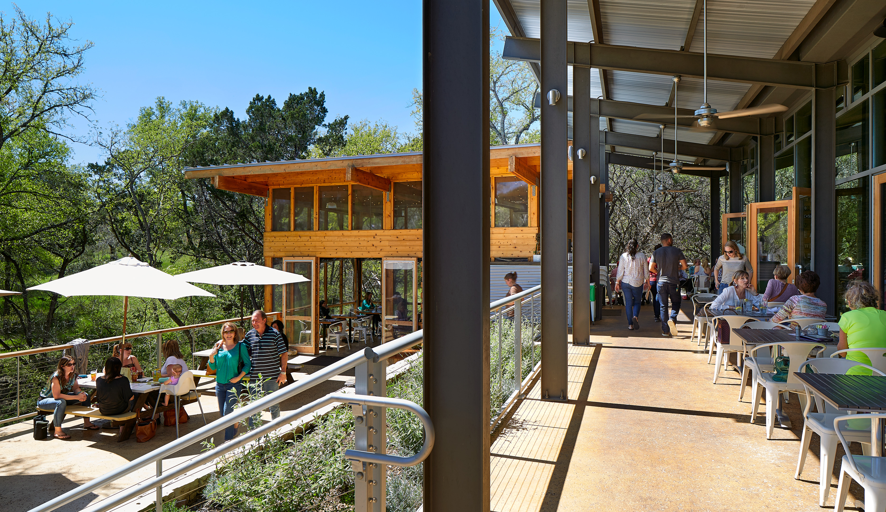 People are dining and socializing on an outdoor patio with tables and umbrellas. The patio, surrounded by greenery and featuring a wooden building, creates a perfect Auto Draft for relaxation.