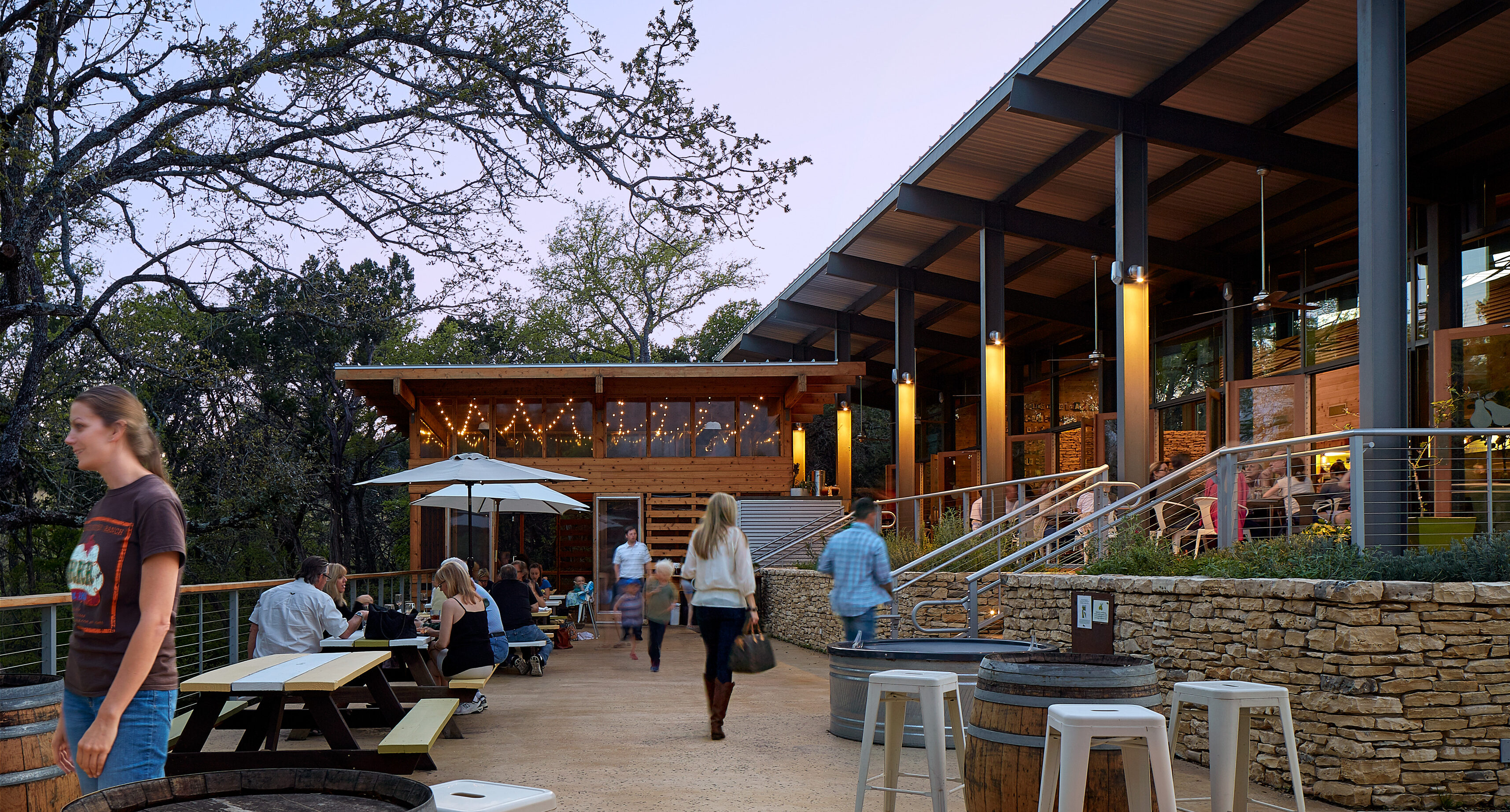 Outdoor seating area of a modern restaurant with people walking and sitting at tables. Stone walls, wooden furniture, and a large canopy are visible, with trees in the background. Evening lighting gives it an ambiance that looks straight out of an Auto Draft design.