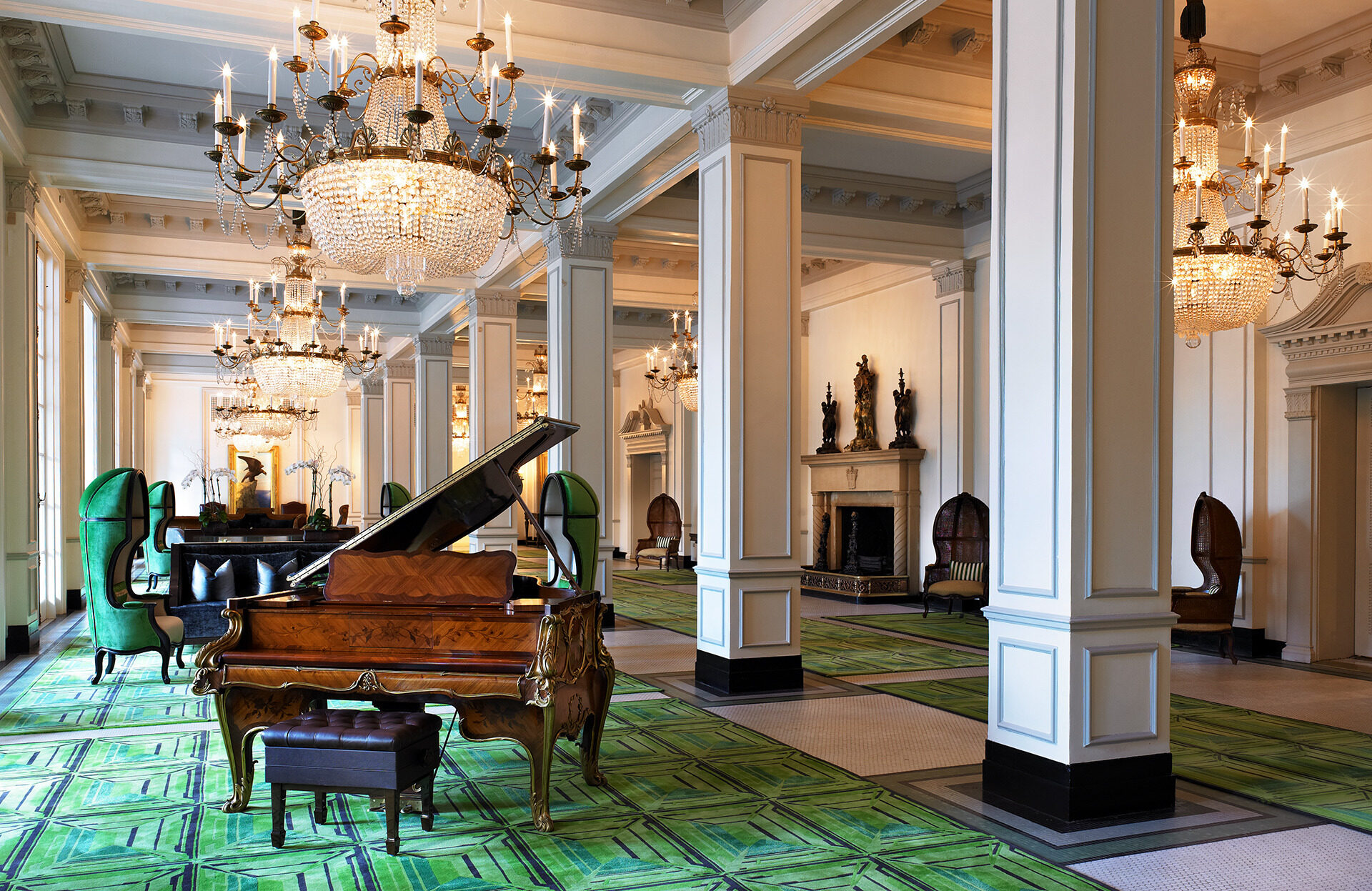 Elegant room with chandeliers, a grand piano, patterned green carpet, and luxurious seating. Pillars and classical architectural details enhance the sophisticated ambiance.