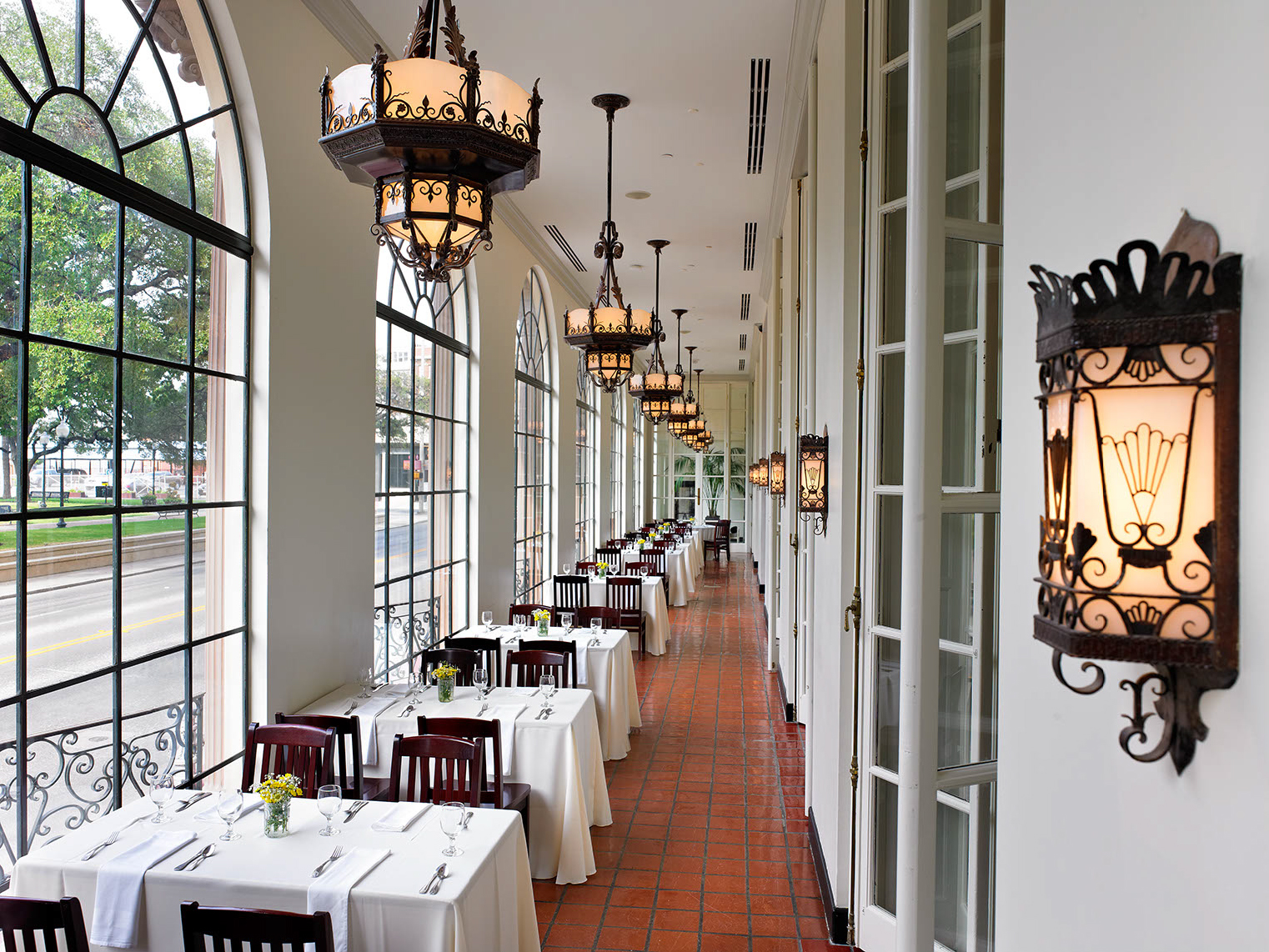 A long, elegant restaurant dining area with arched windows, white tablecloths, dark wooden chairs, and vintage chandeliers.