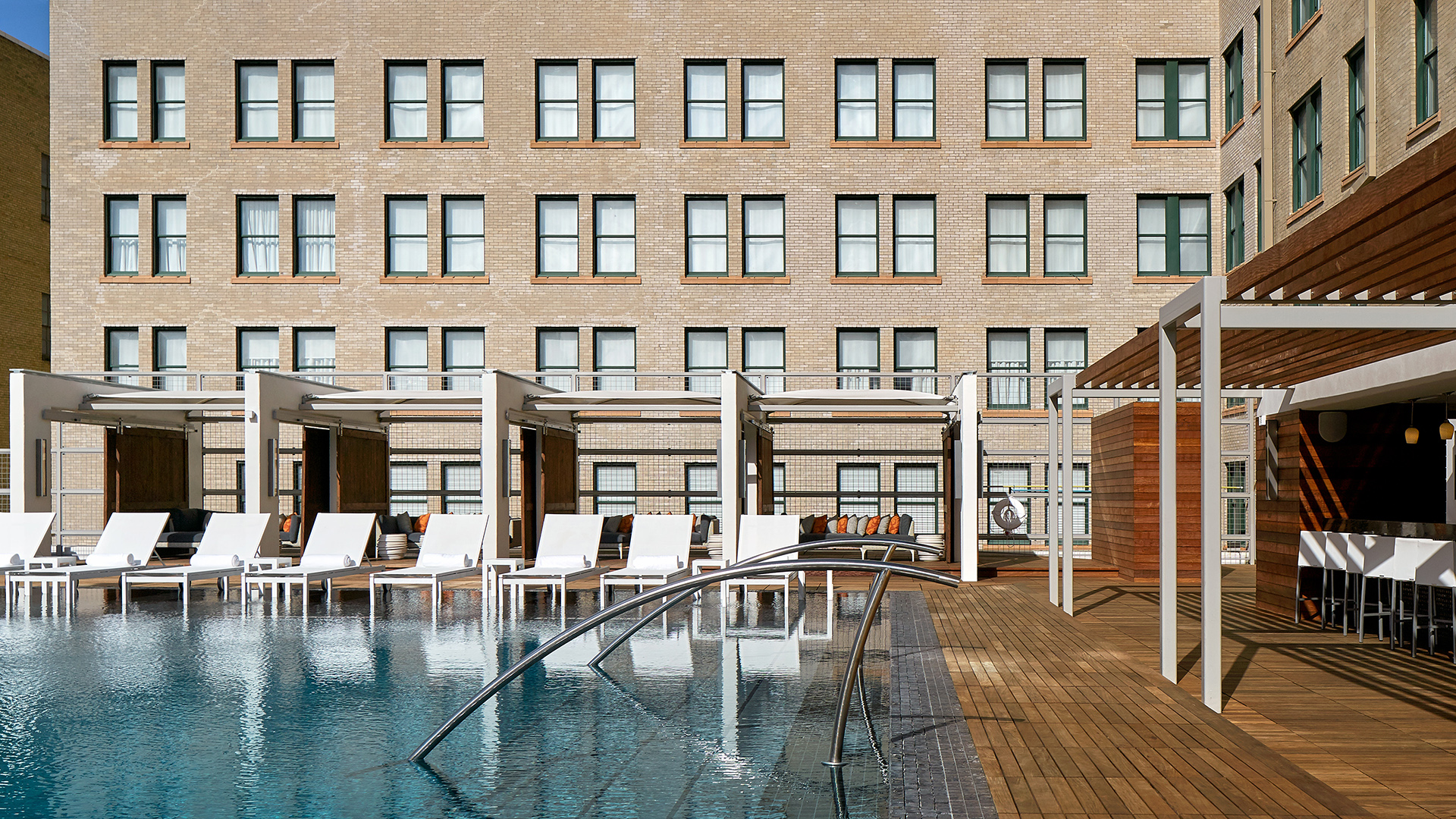 Outdoor pool area with white lounge chairs and cabanas on a wooden deck, adjacent to a multi-story brick building with numerous windows.