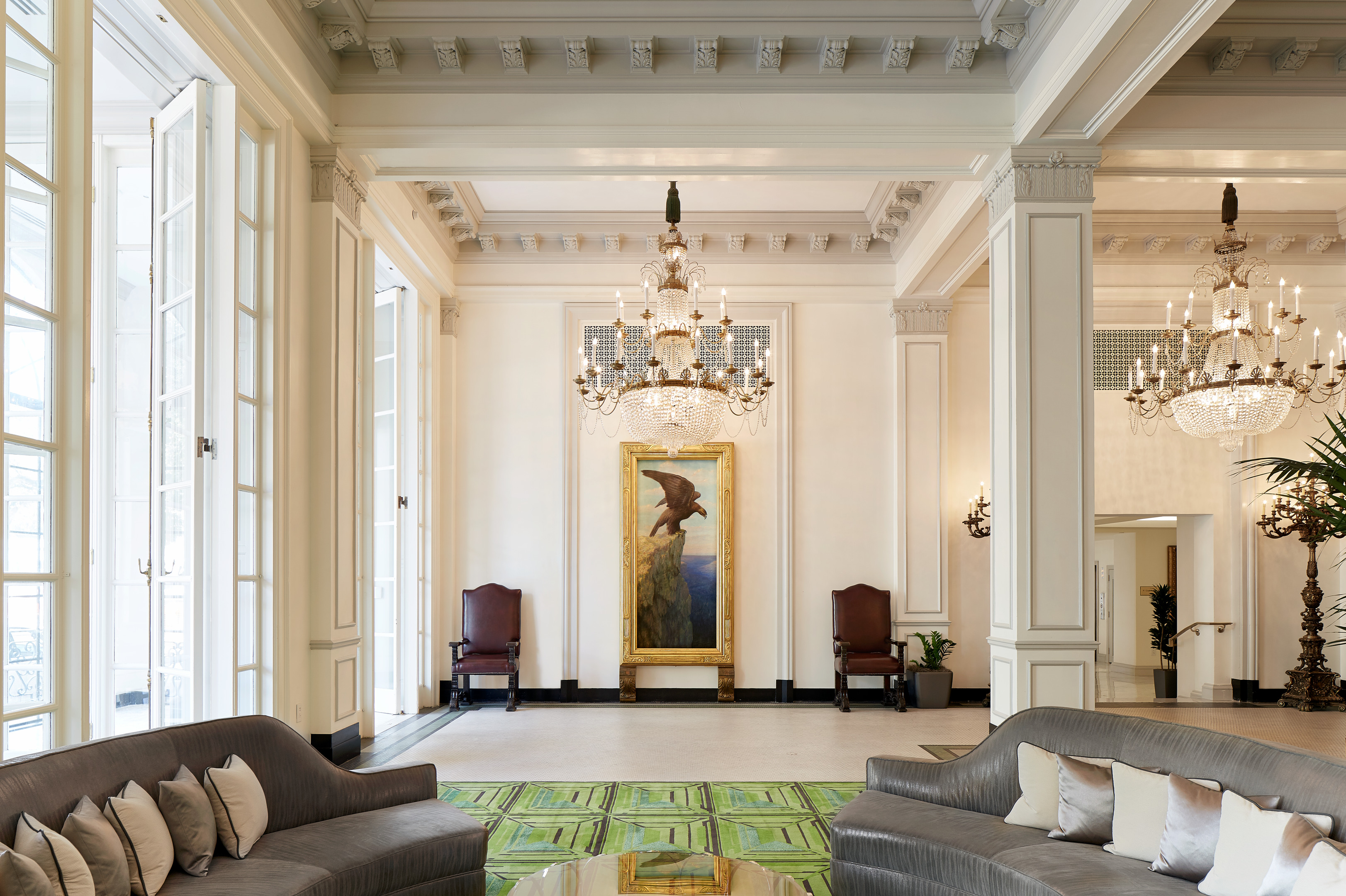 Elegant hotel lobby with high ceilings, chandeliers, large windows, and a central painting. Gray sofas and green carpet add to the stylish decor.