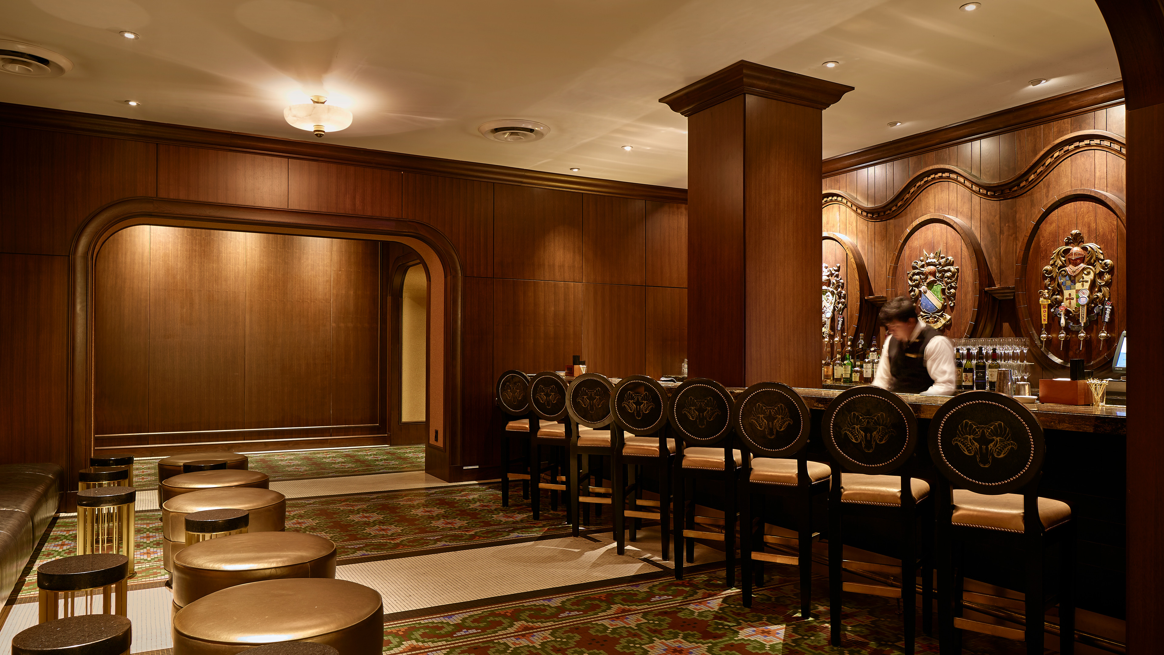 A dimly-lit bar with ornate wood paneling. A bartender is preparing drinks behind the counter, and high-backed bar stools with intricate designs line the bar. The seating area has additional small round tables and stools.