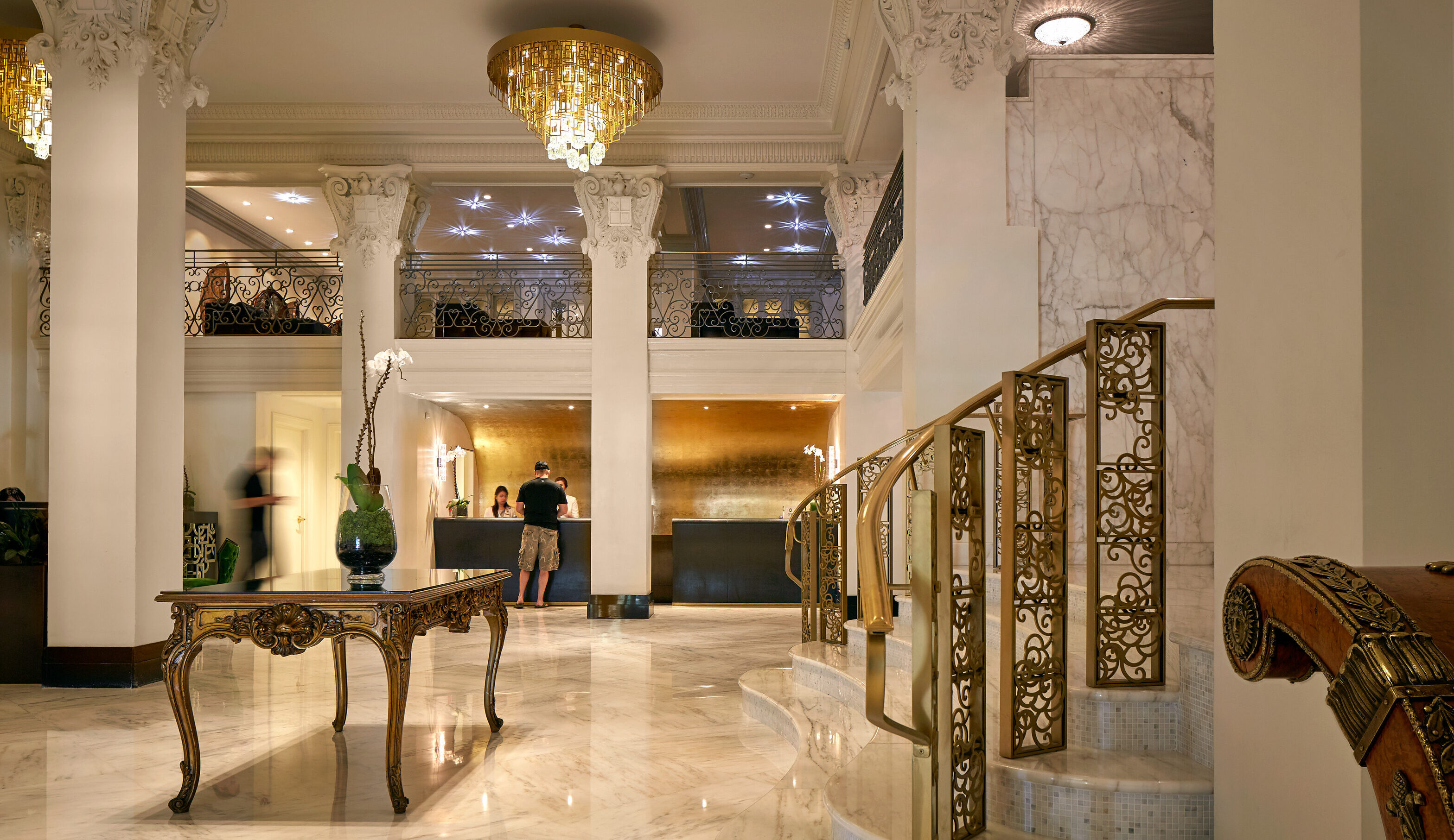 A luxurious hotel lobby with marble floors, ornate columns, a grand staircase, and a chandelier. Guests interact at the reception desk in the distance.