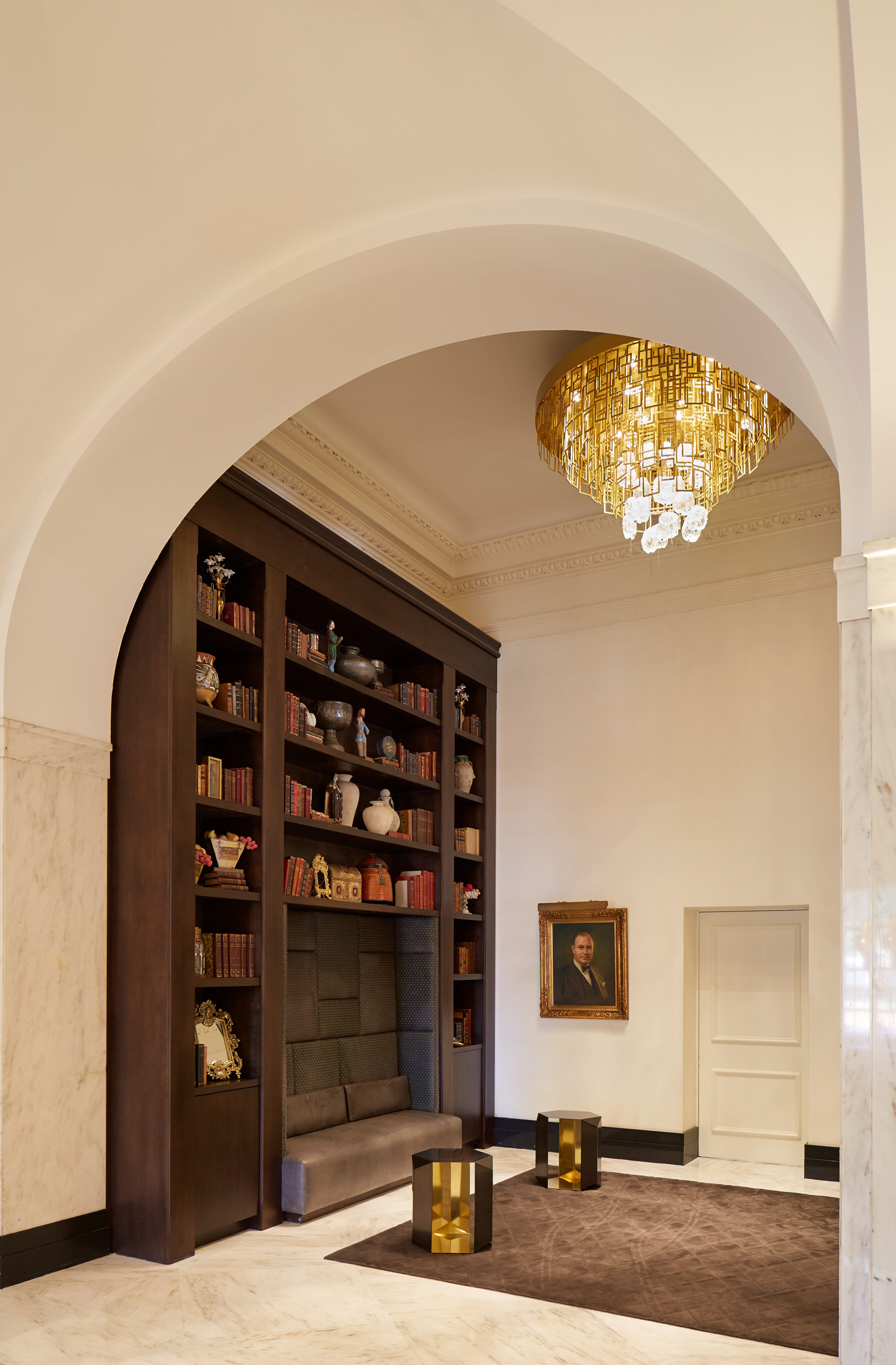 A luxurious interior features an arched doorway, a chandelier, dark wood bookshelves with decorative items, a built-in cushioned seat, a portrait on the wall, and two modern stools on a brown rug.