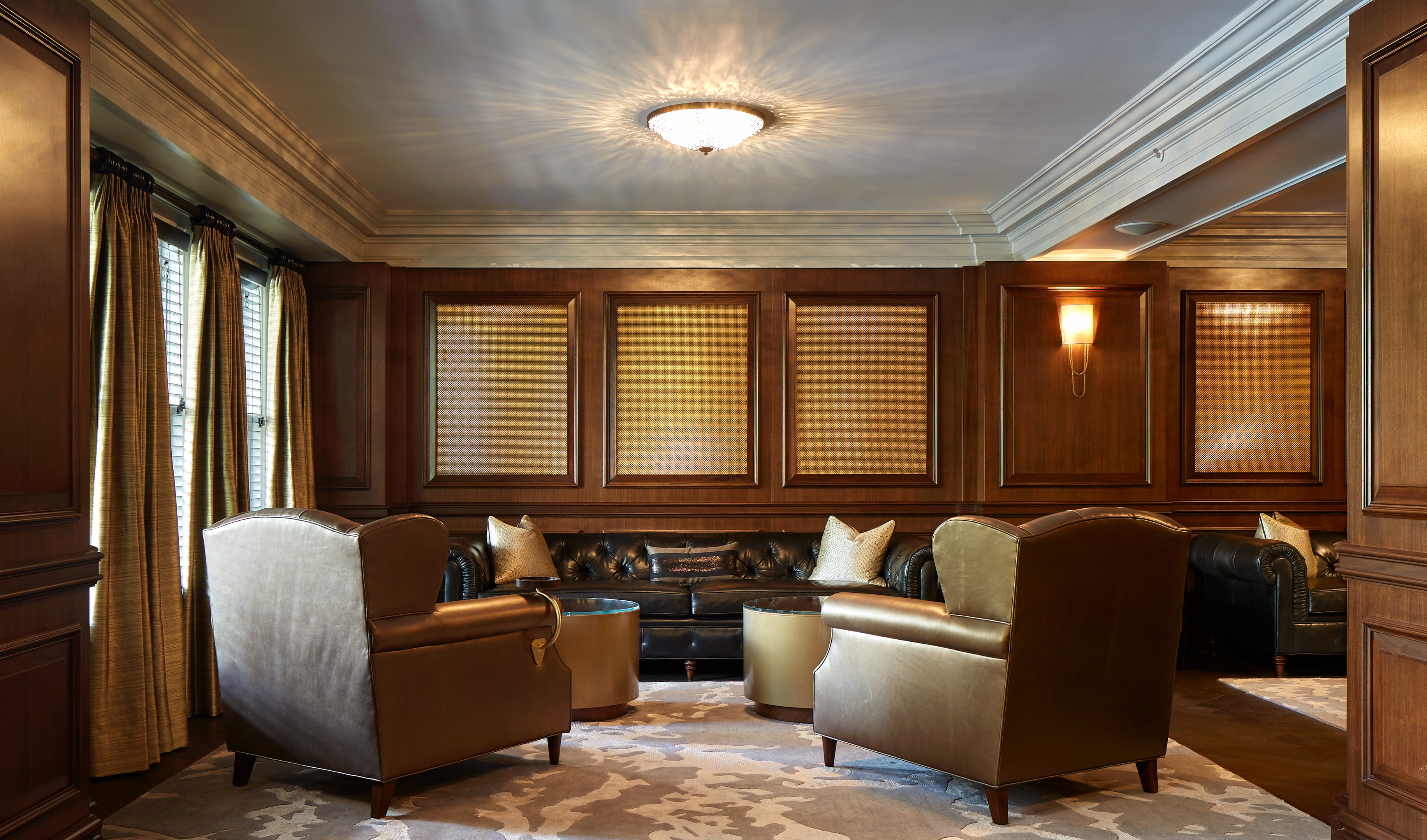 A warmly lit, wood-paneled room with two leather armchairs, a black sofa, and round coffee tables. Covered windows and a patterned rug complete the cozy setup, reminiscent of a suite at the luxury St. Anthony hotel.