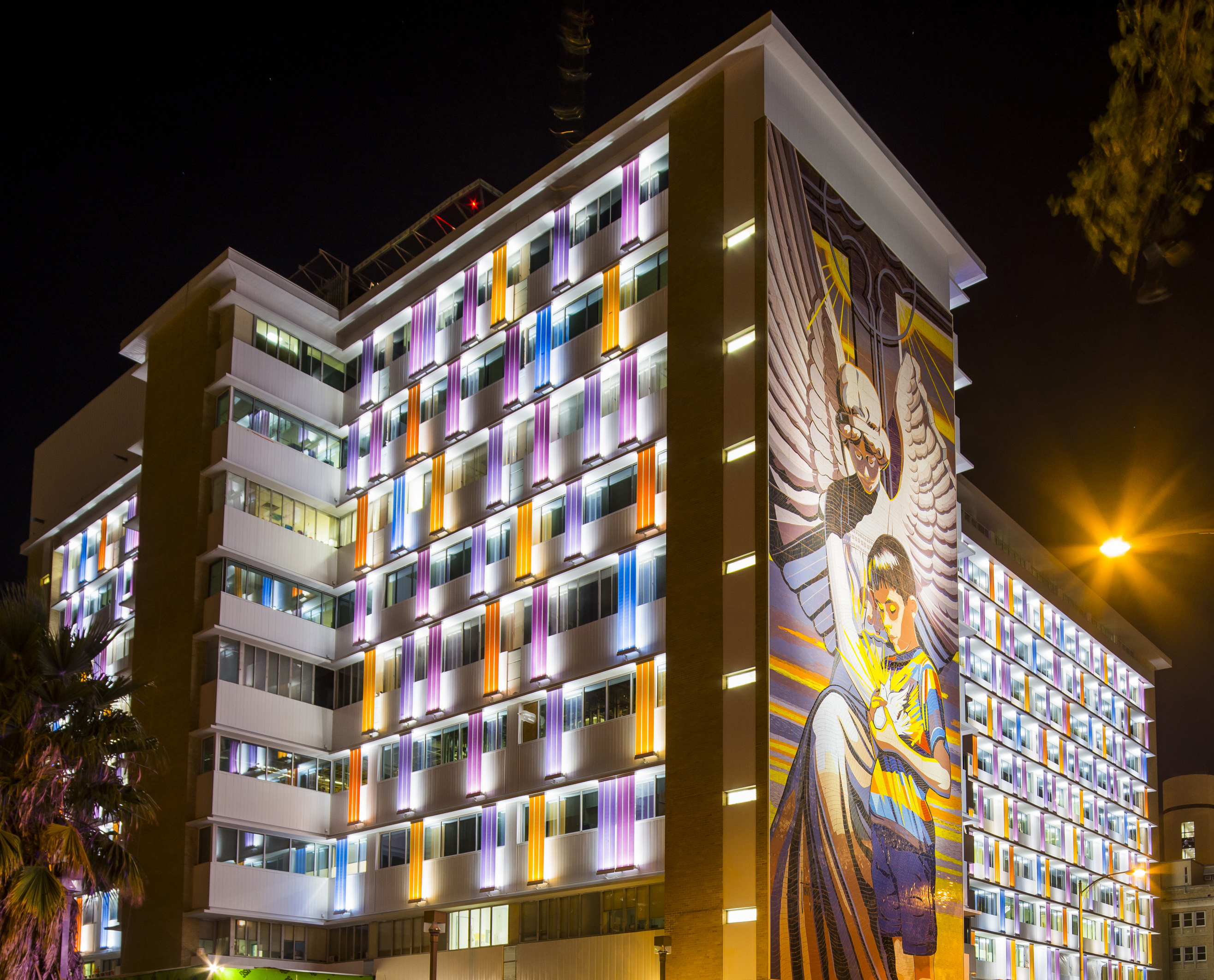 A multi-story building at night is illuminated with colorful lights and features a large mural of a winged figure and a person on its facade, resembling the vibrant spirit of CHRISTUS Children's Hospital in San Antonio.