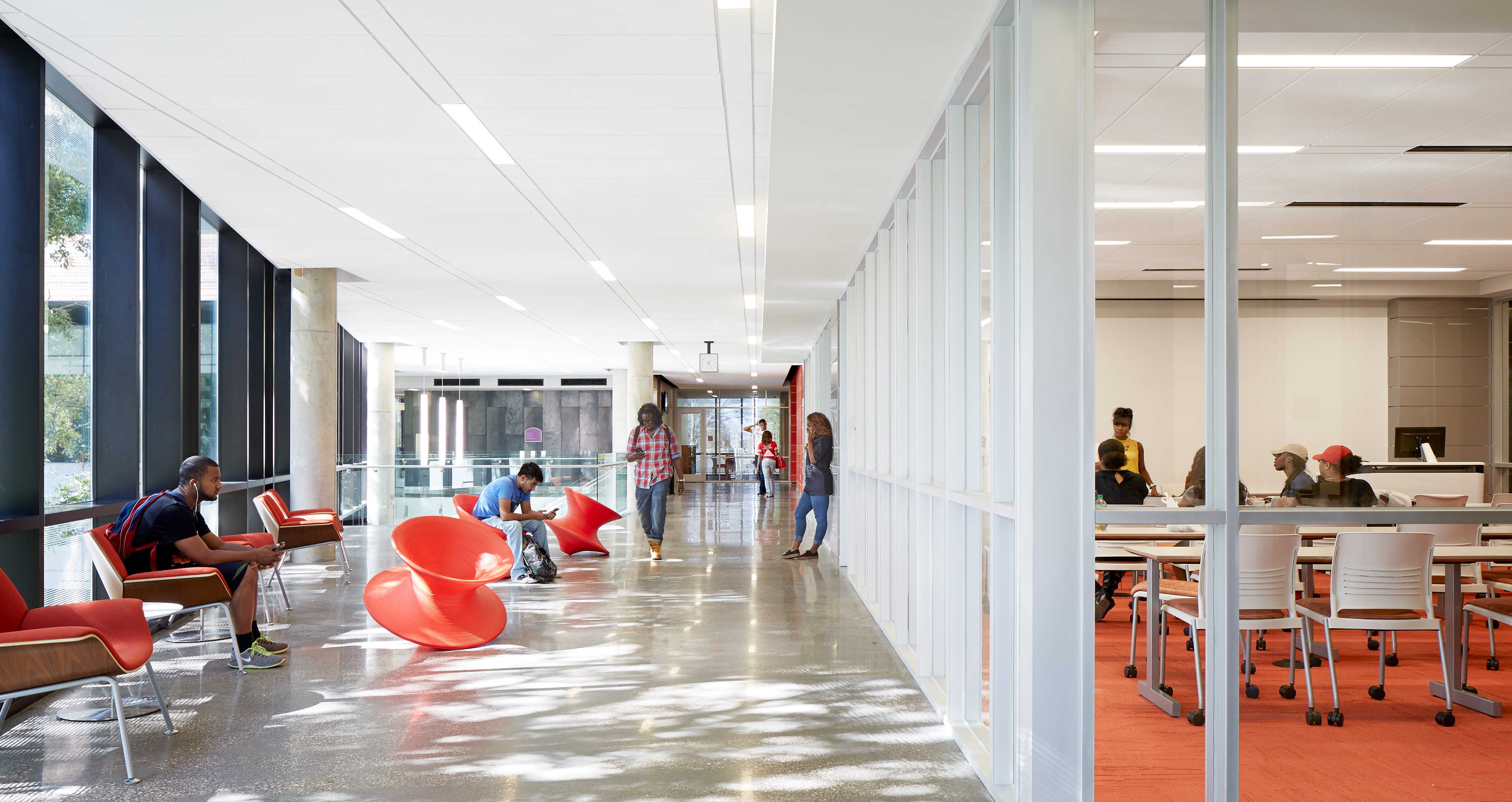 Bright hallway in a modern building with people seated and standing, featuring red swivel chairs and glass-walled rooms.