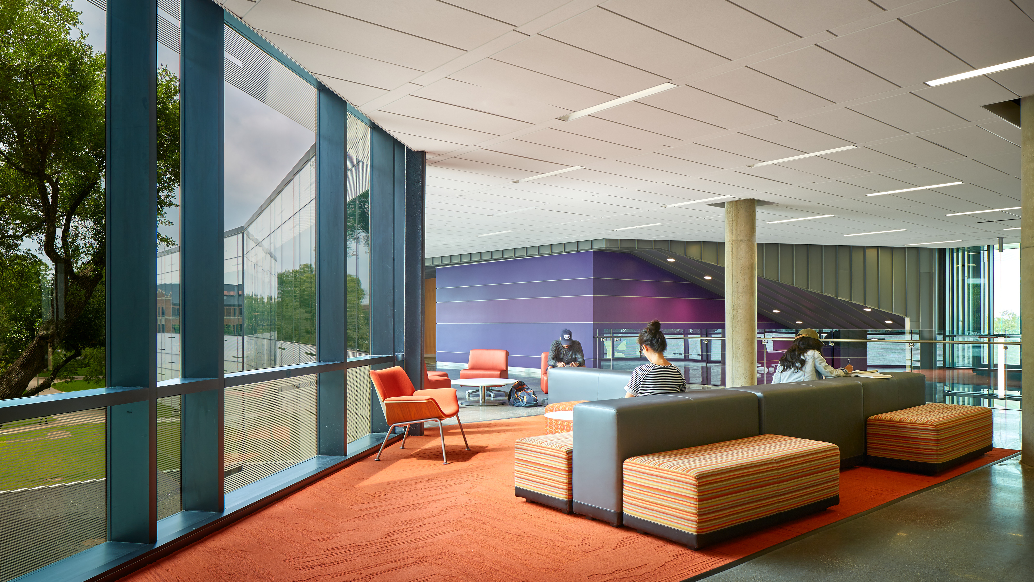 A modern lounge area with large windows, orange and gray furniture, a purple wall, and people seated and working on laptops.