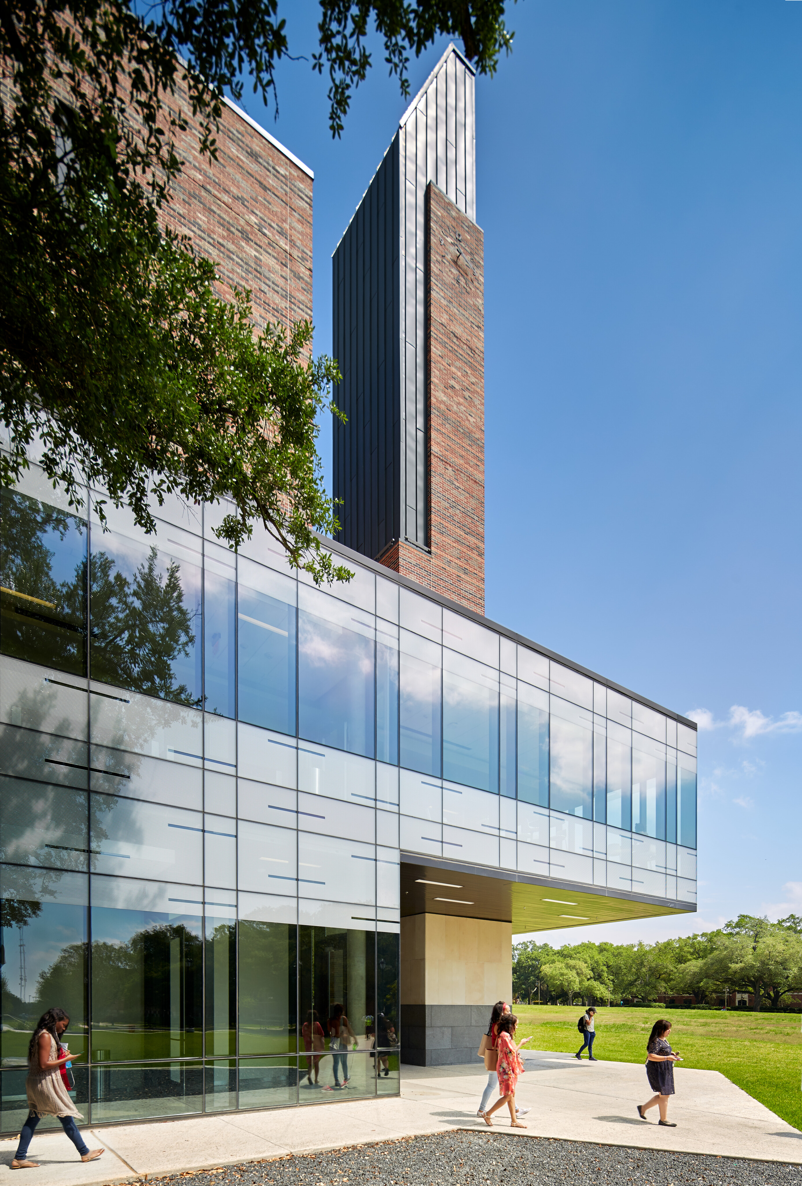 A modern building with large glass windows and two tall towers. People walk along the pathway beside the building on a sunny day, with green grass and trees in the background.