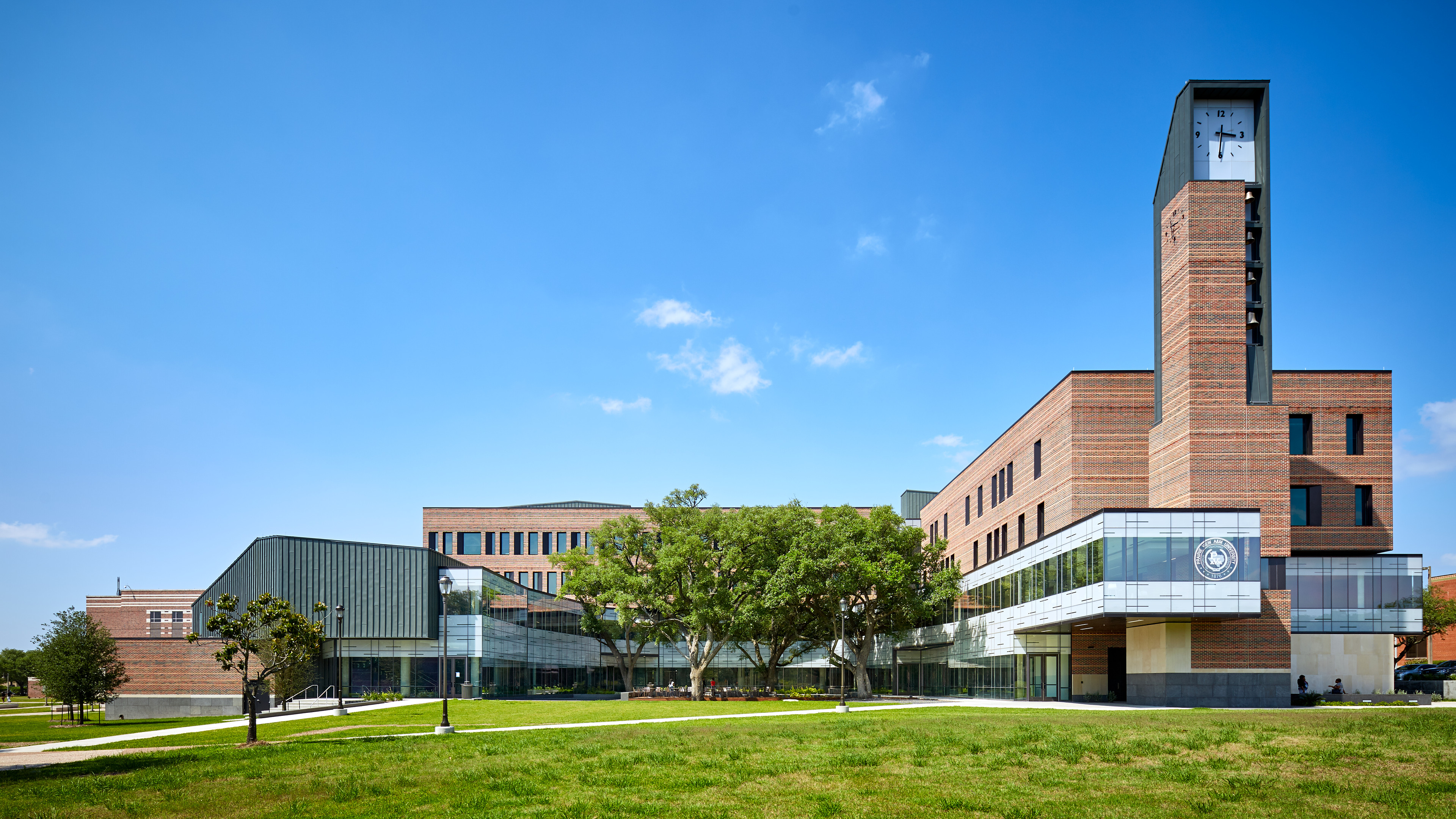 A modern brick university building with a clock tower and large windows under a blue sky. Trees and a grassy lawn are in the foreground.