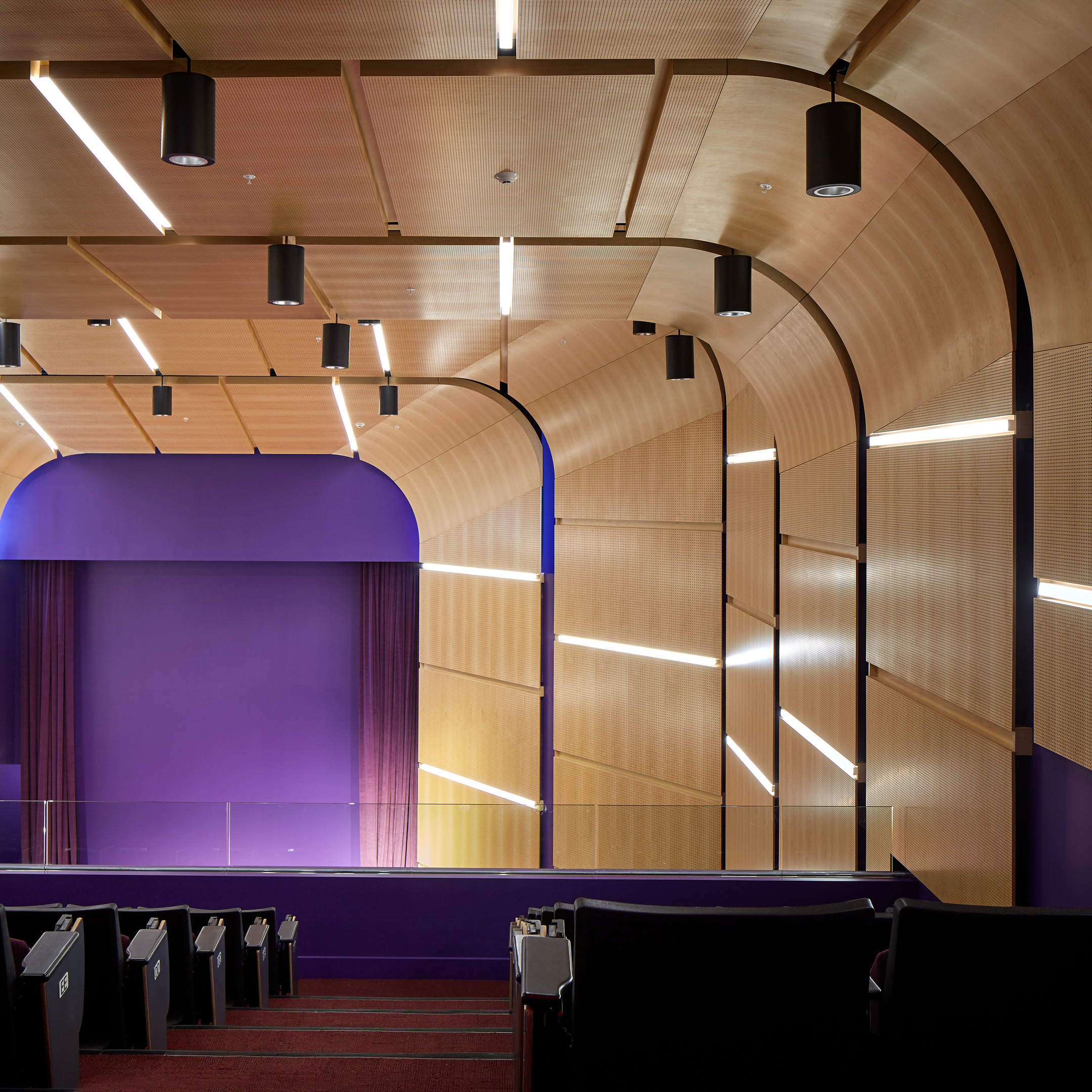 The PrairieView A&M Multipurpose Building boasts a modern auditorium with curved wooden walls, purple curtains, and evenly spaced linear lighting. Rows of seating are visible in the foreground, all facing the stage.