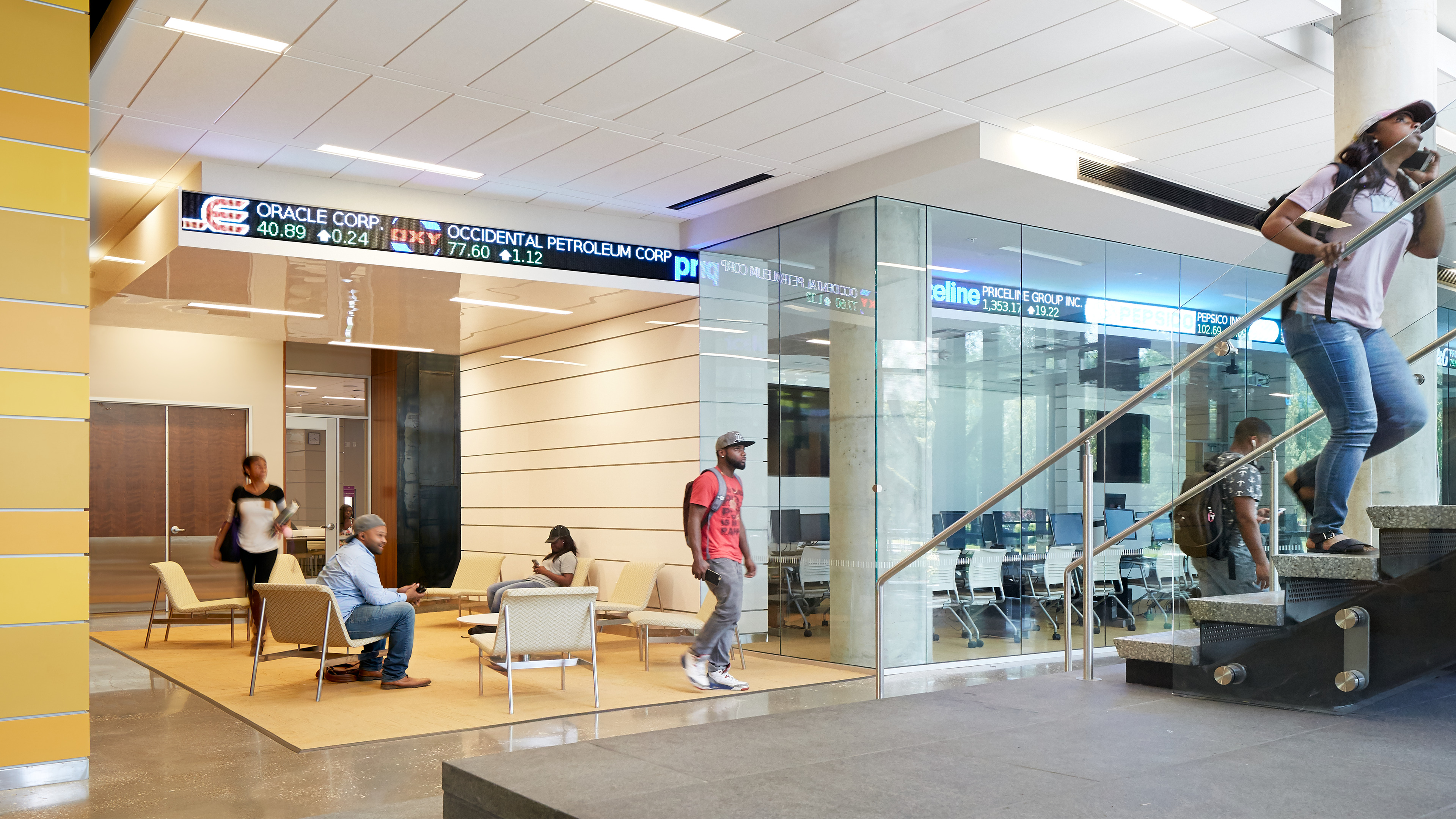 A modern lobby area with several people seated, standing, and walking. A stock ticker is displayed on the upper wall, and a person is seen going up the stairs. Glass-walled rooms in the background.