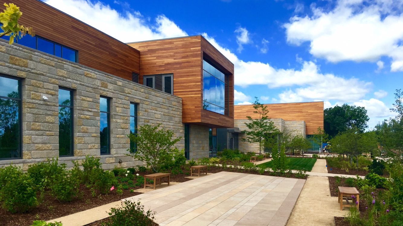 Modern building with stone and wood facade, large windows, and landscaped courtyard featuring benches and pathways under a blue sky with scattered clouds.