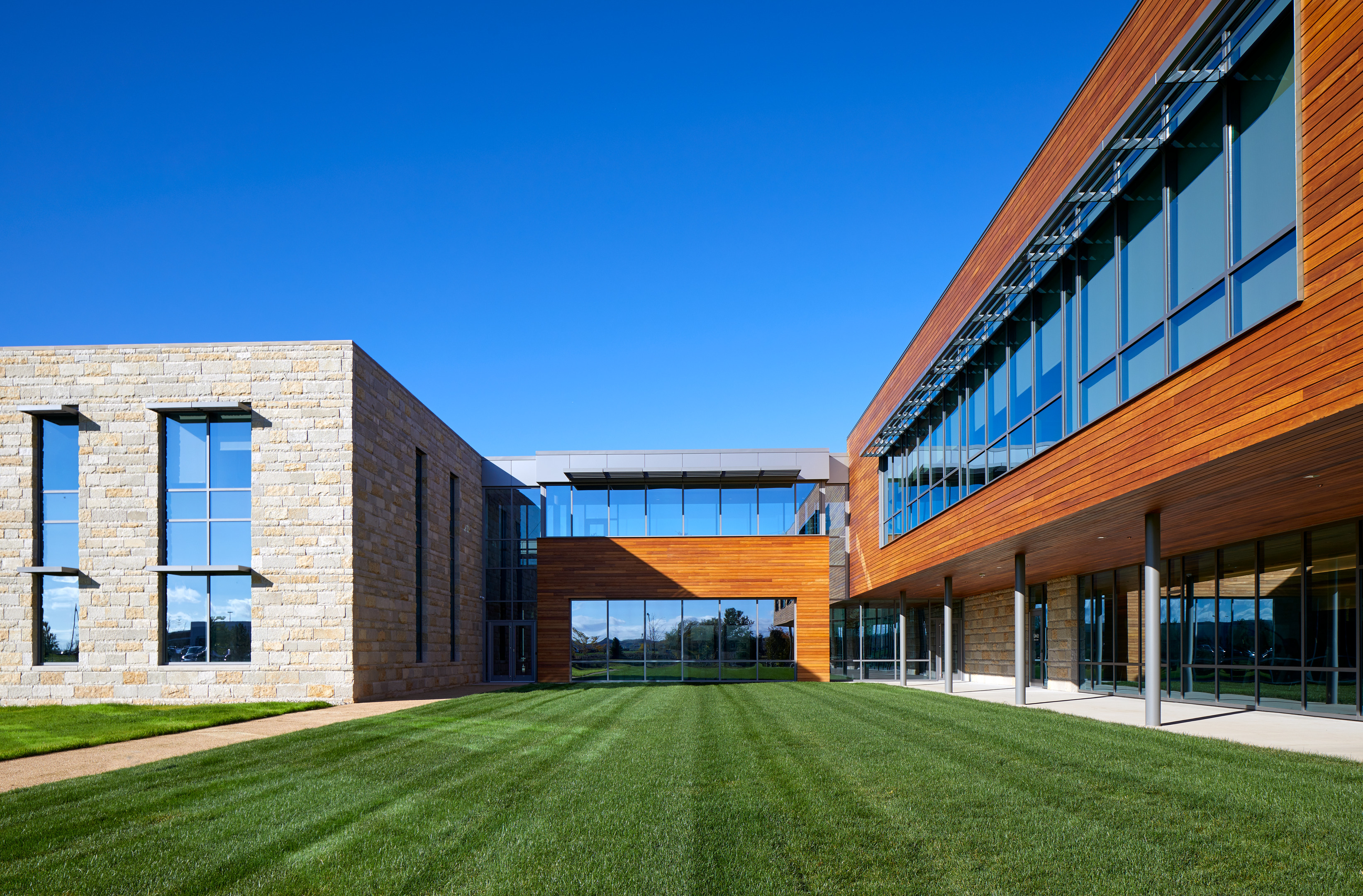 Modern building with a mix of stone and wooden facades, featuring large glass windows and a well-manicured grassy courtyard under a clear blue sky.