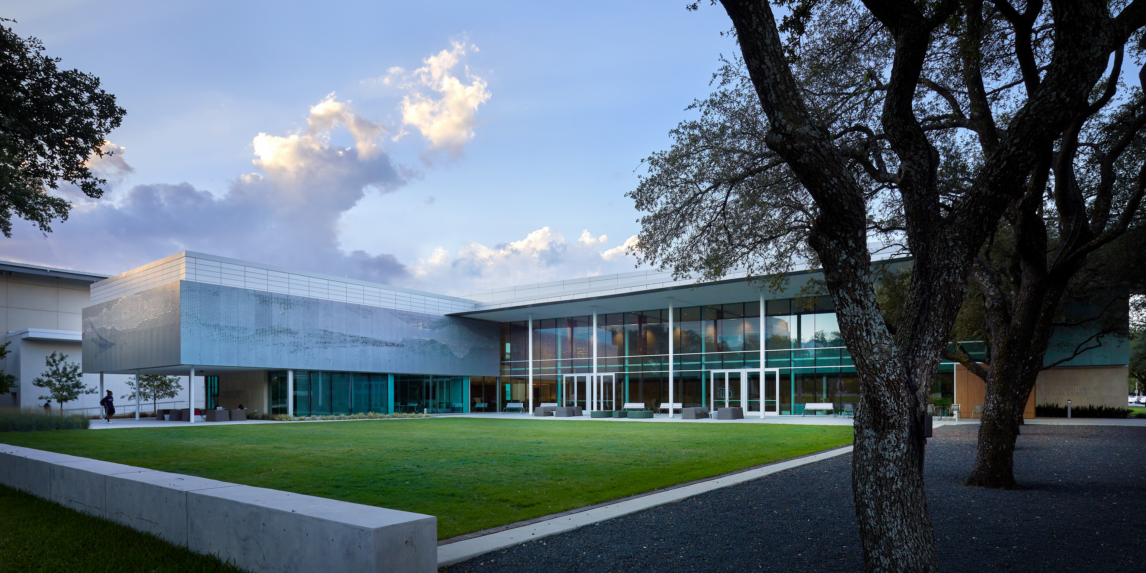 Modern building with large windows and glass walls, surrounded by trees and a green lawn, photographed under a partly cloudy sky. This stunning structure serves as the University of Texas Alumni Center in Dallas, blending contemporary design with natural beauty.