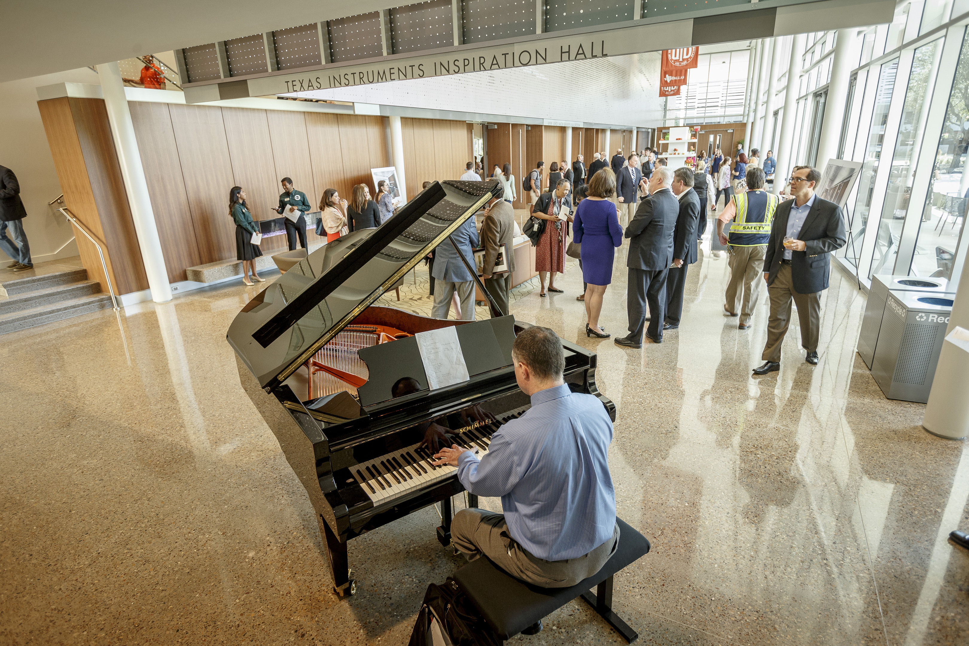 An individual plays a grand piano in a hall where people are conversing and networking. The space, marked "Texas Instruments Inspiration Hall," is part of the Alumni Center at the University of Texas in Dallas.