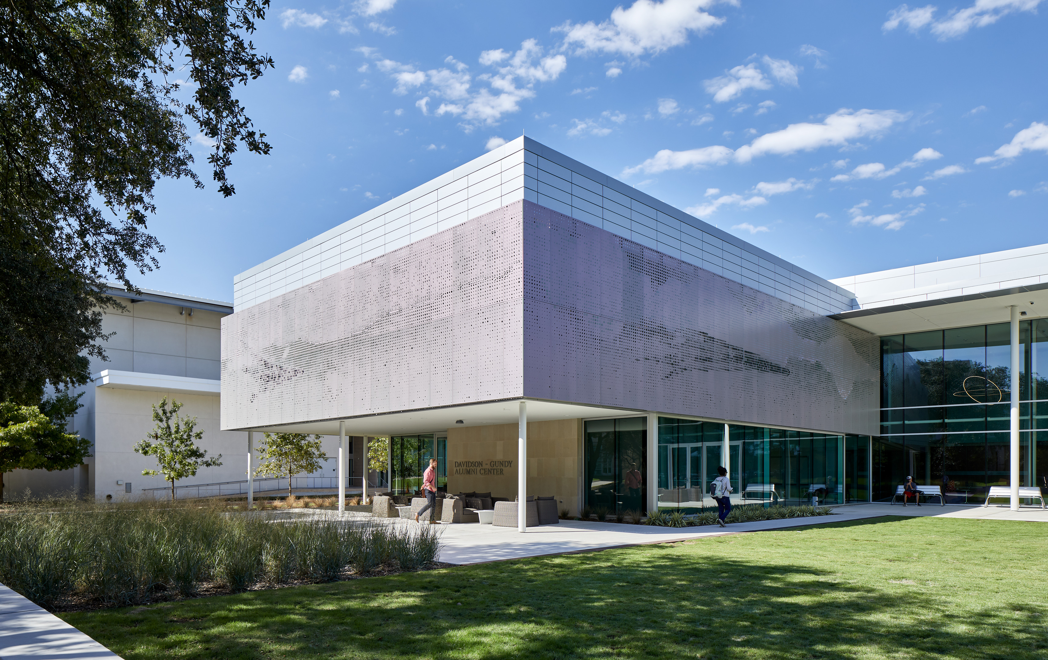 A modern building with a large, reflective metal facade, surrounded by a well-maintained lawn and trees, under a clear sky with scattered clouds. People are walking near the entrance.