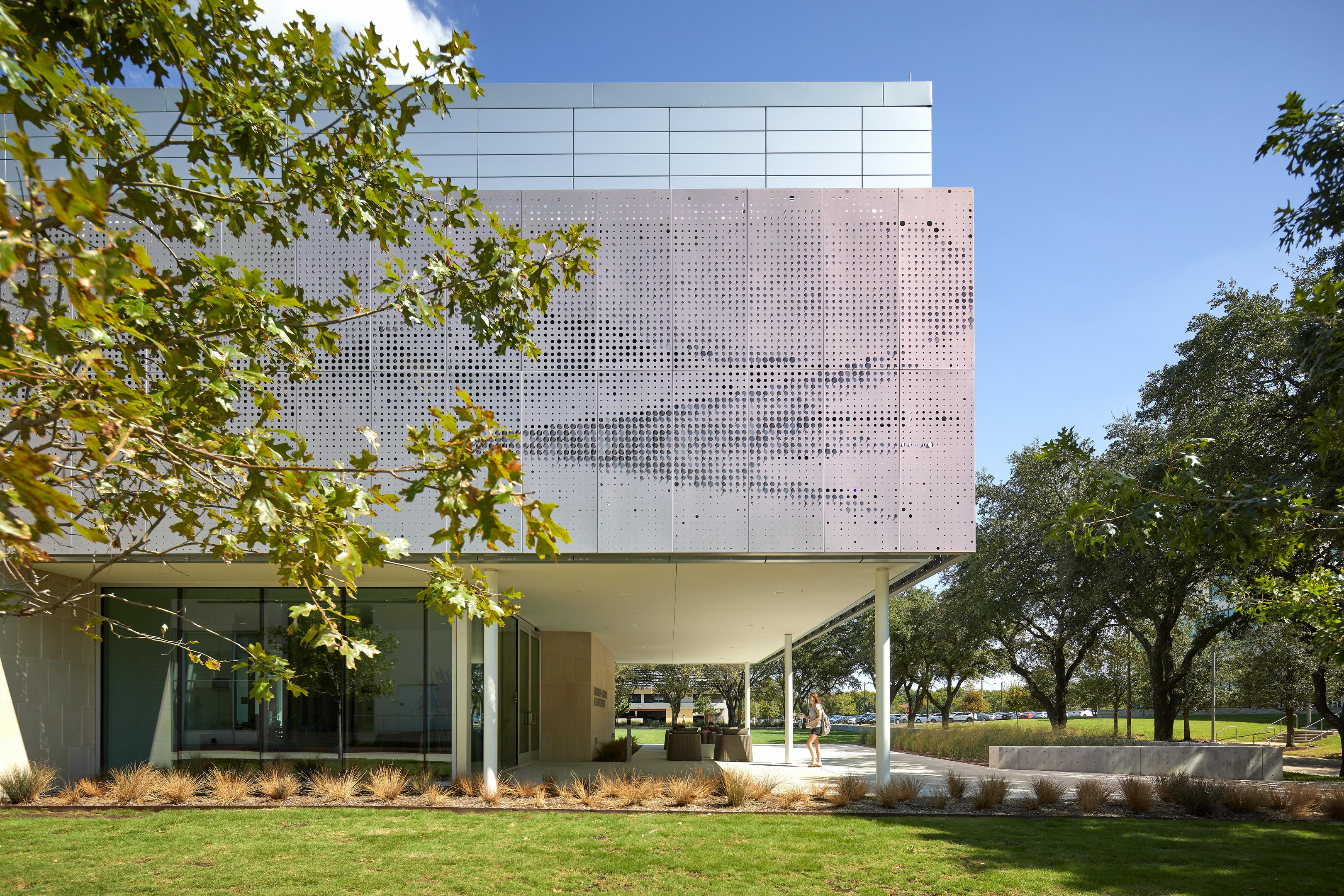 A modern building with perforated metal facade, large glass windows, and a grassy lawn with trees on a clear day.