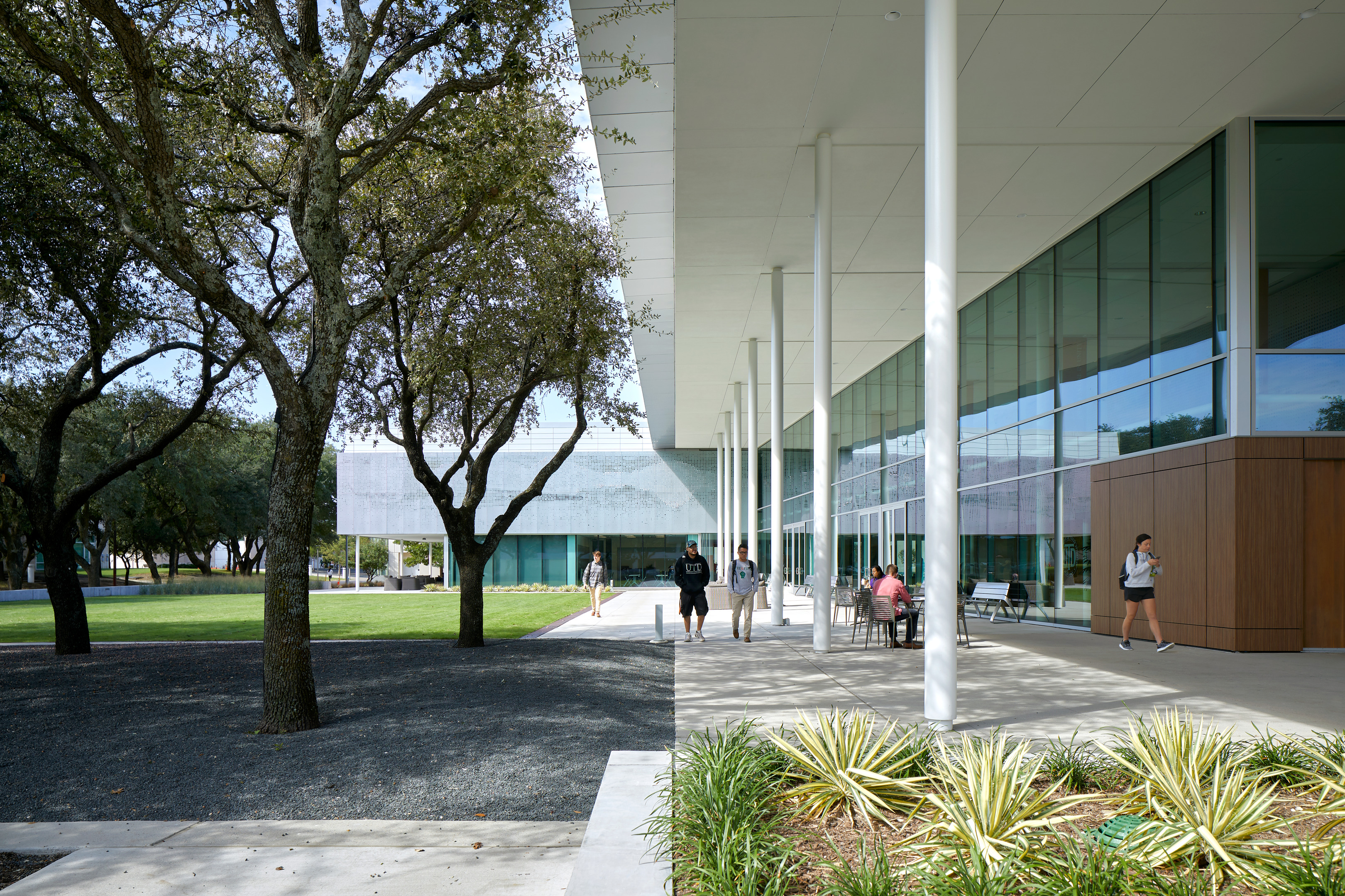 Modern building with large glass windows and a covered walkway, surrounded by trees and landscaped greenery. Several people walk and sit along the pathway.