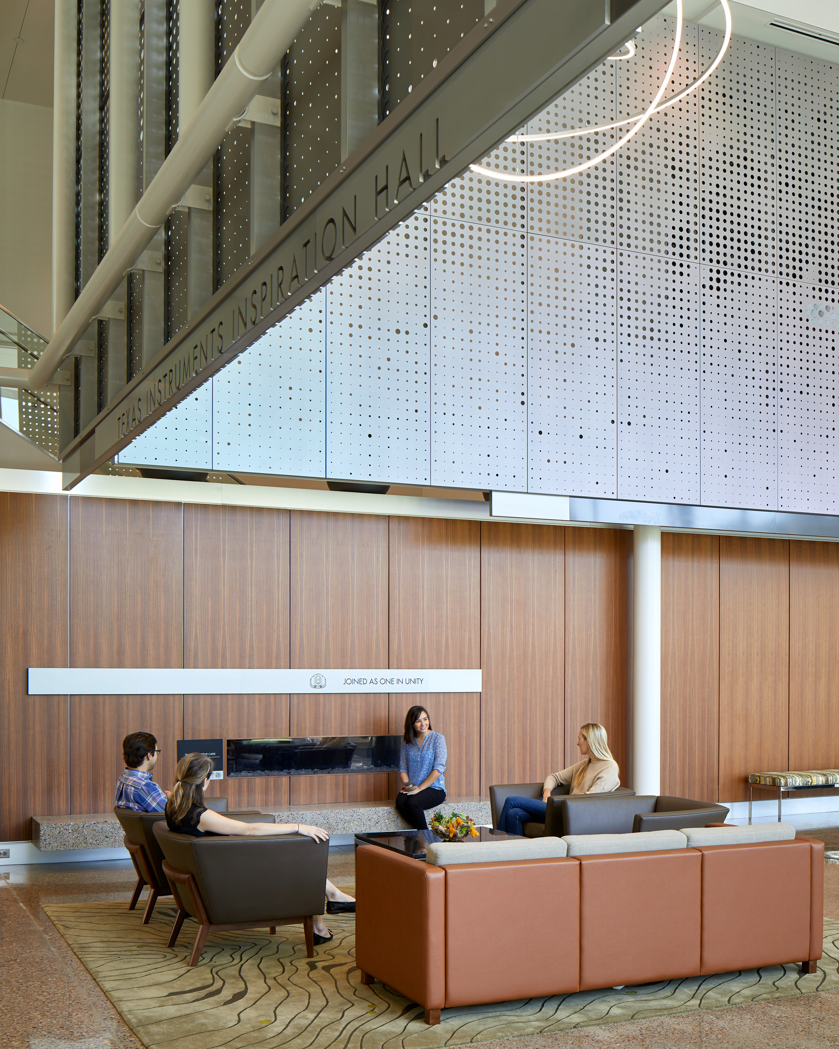 Four people are seated on couches and chairs in a modern lounge area under a sign that reads "Texas Instruments Inspiration Hall.