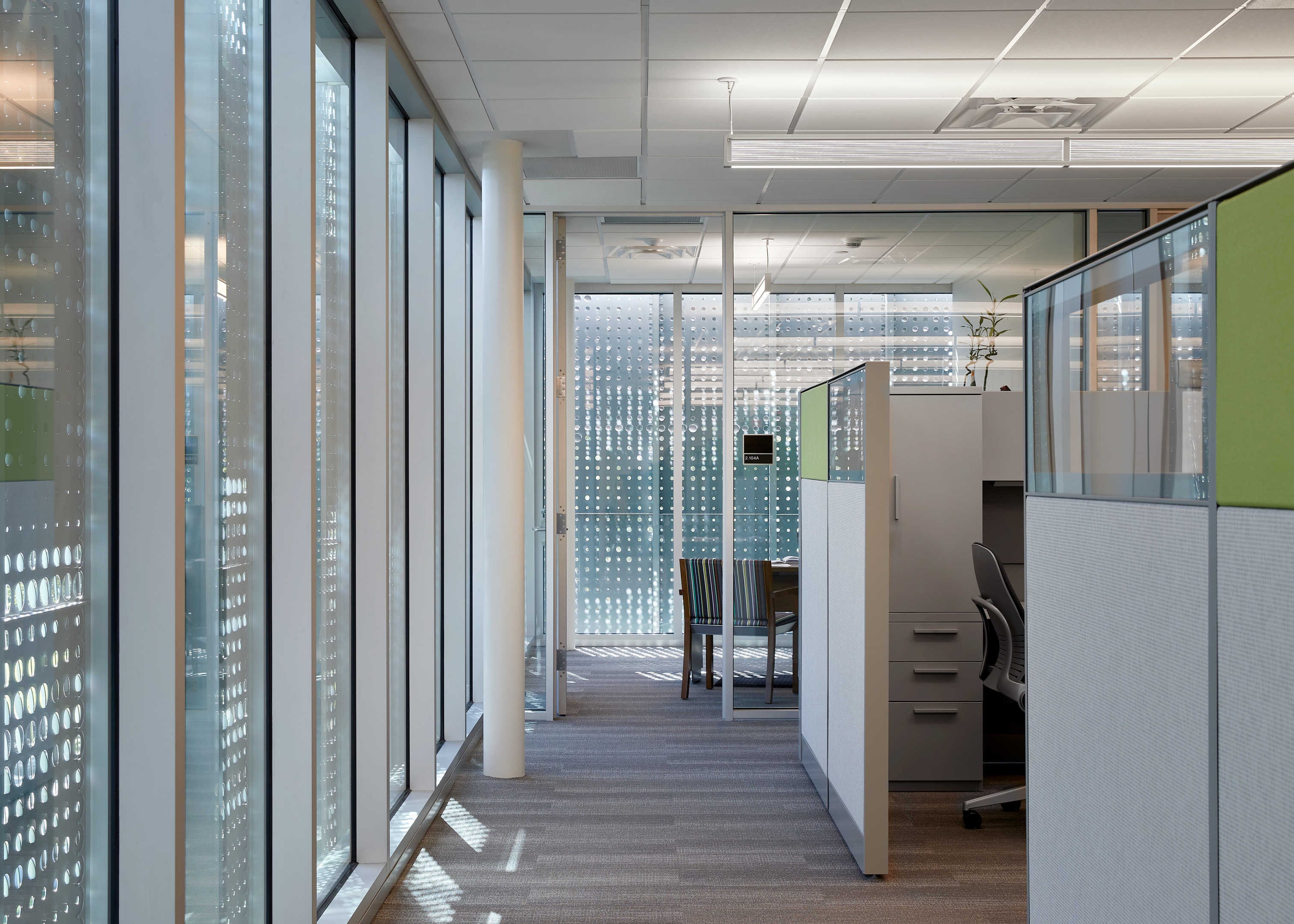 A modern office space with large windows, cubicles, and a conference room. The area is well-lit, with patterned glass walls and a clean, organized arrangement.