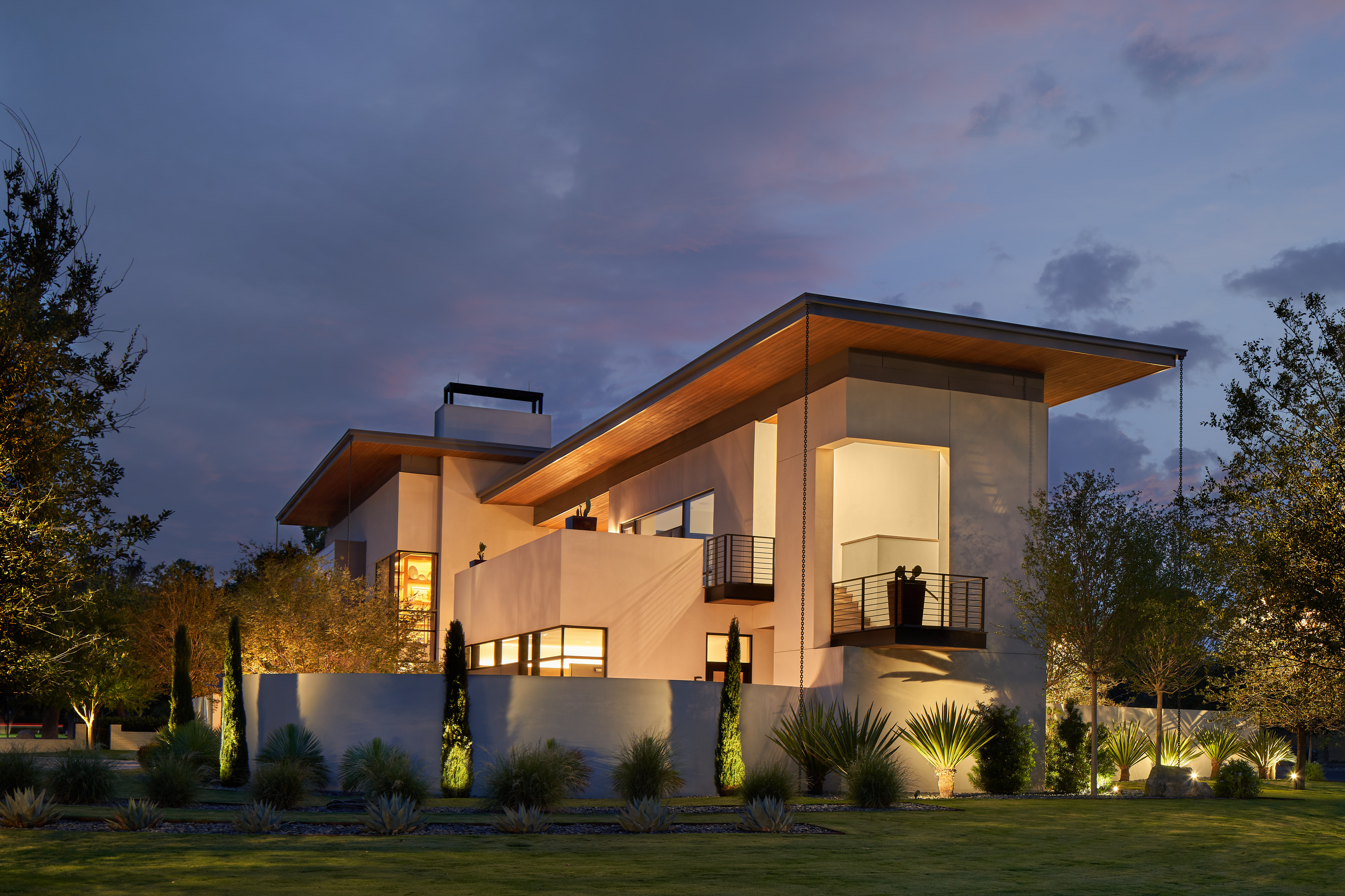 The Midland Residence is a modern two-story house with large windows and lights illuminating the exterior at dusk, surrounded by landscaped gardens and trees.