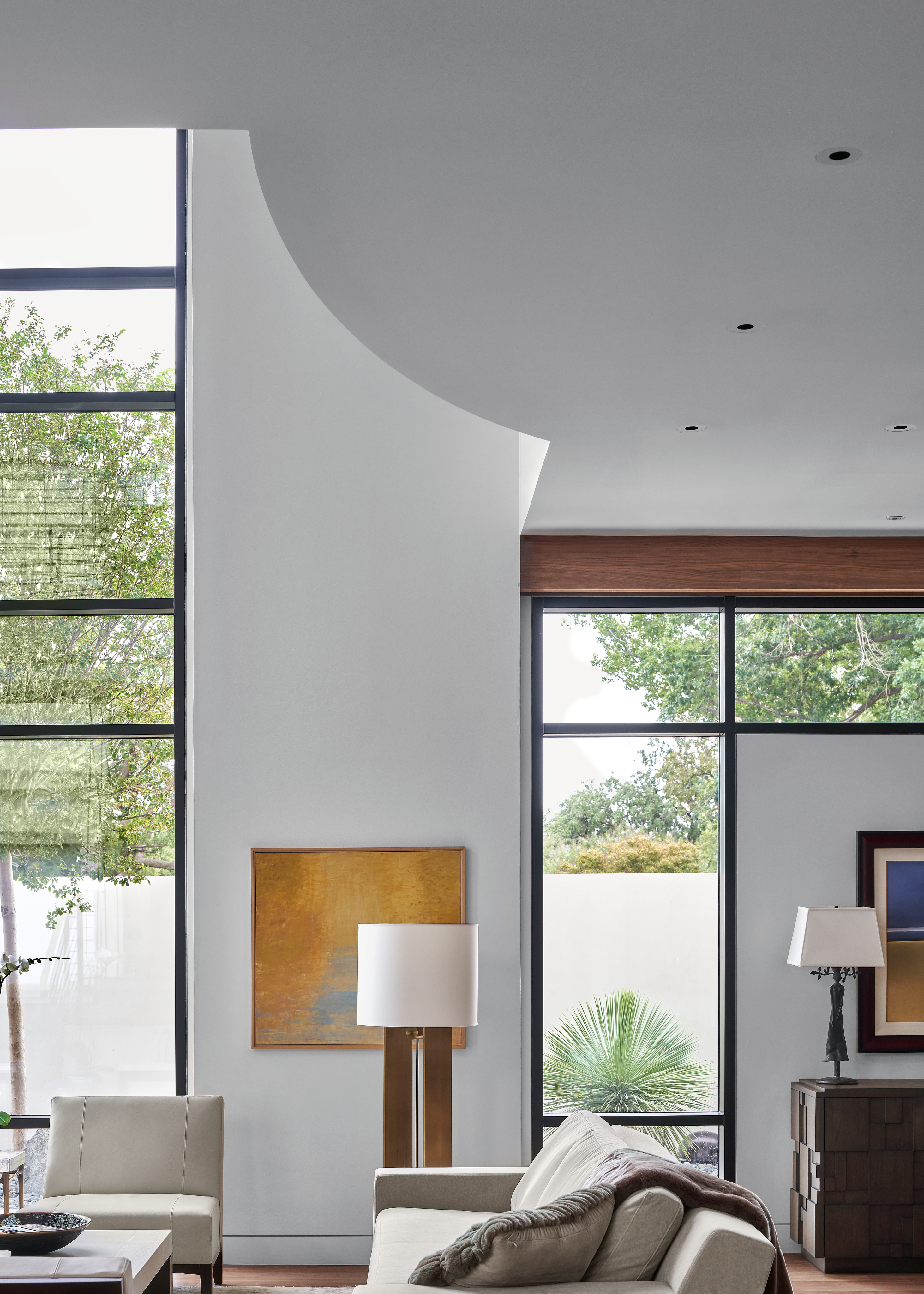 Modern living room with tall windows, a white sofa, wooden furniture, and two lamps. A large abstract painting adorns the wall while trees are visible outside. This stunning space exemplifies the elegant design found in Midland residences.