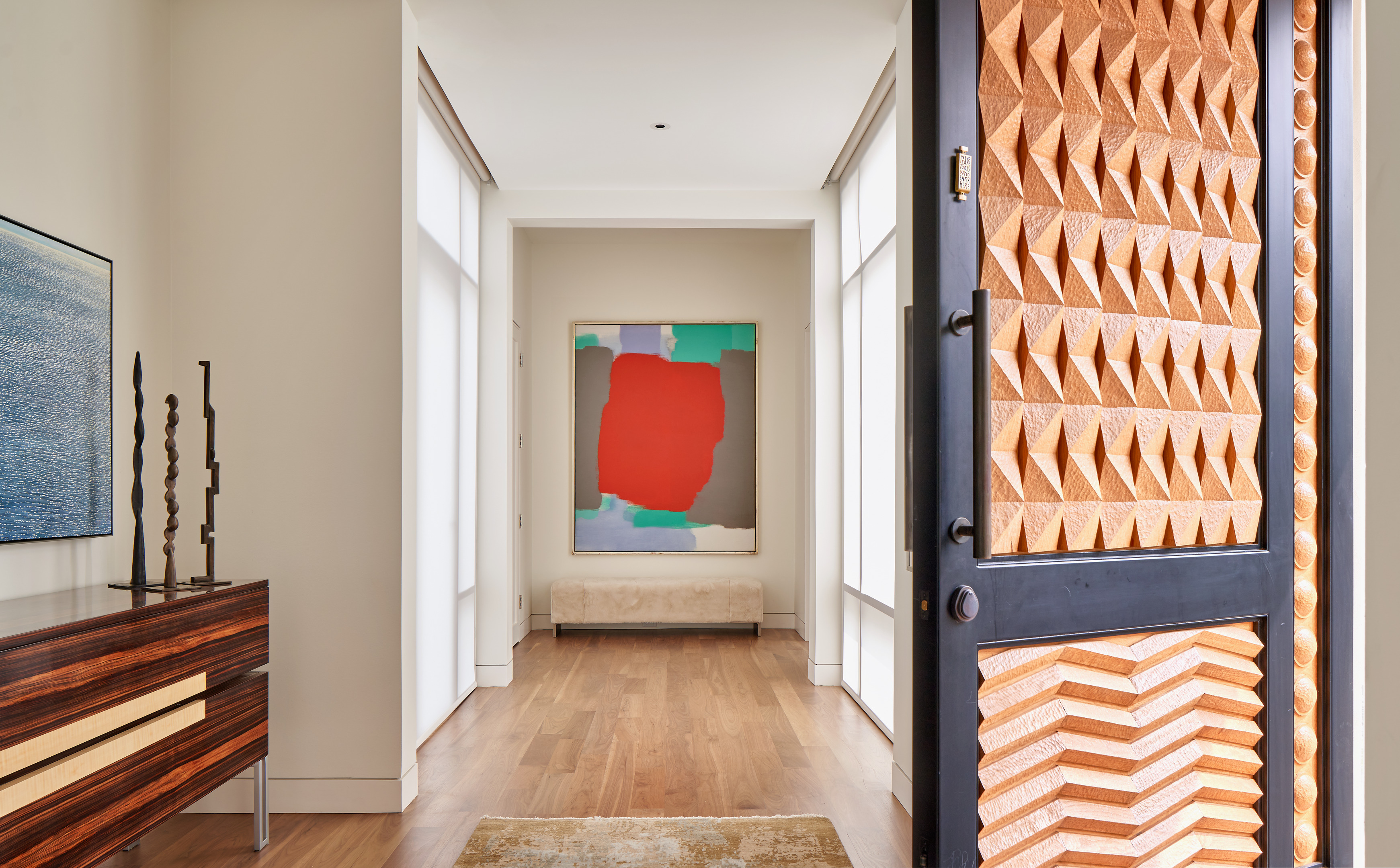 An open door reveals the Midland Residence's modern interior hallway with wooden flooring, a large colorful abstract painting on the far wall, and a contemporary sideboard adorned with decorative items on the left.