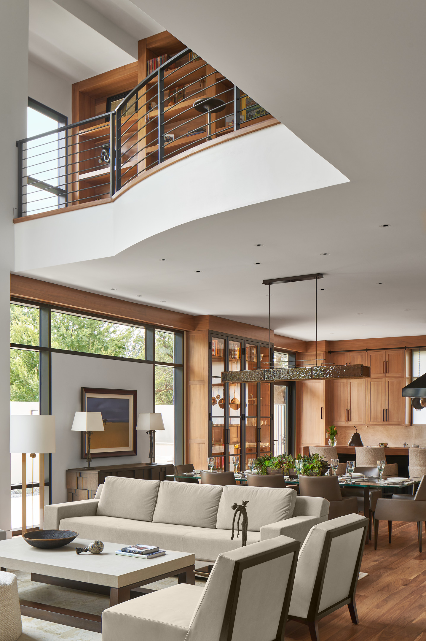 The Midland Residence boasts a modern living and dining area with neutral-tone furniture, extensive wood finishes, large windows, and a mezzanine adorned with bookshelves and a sleek black railing.