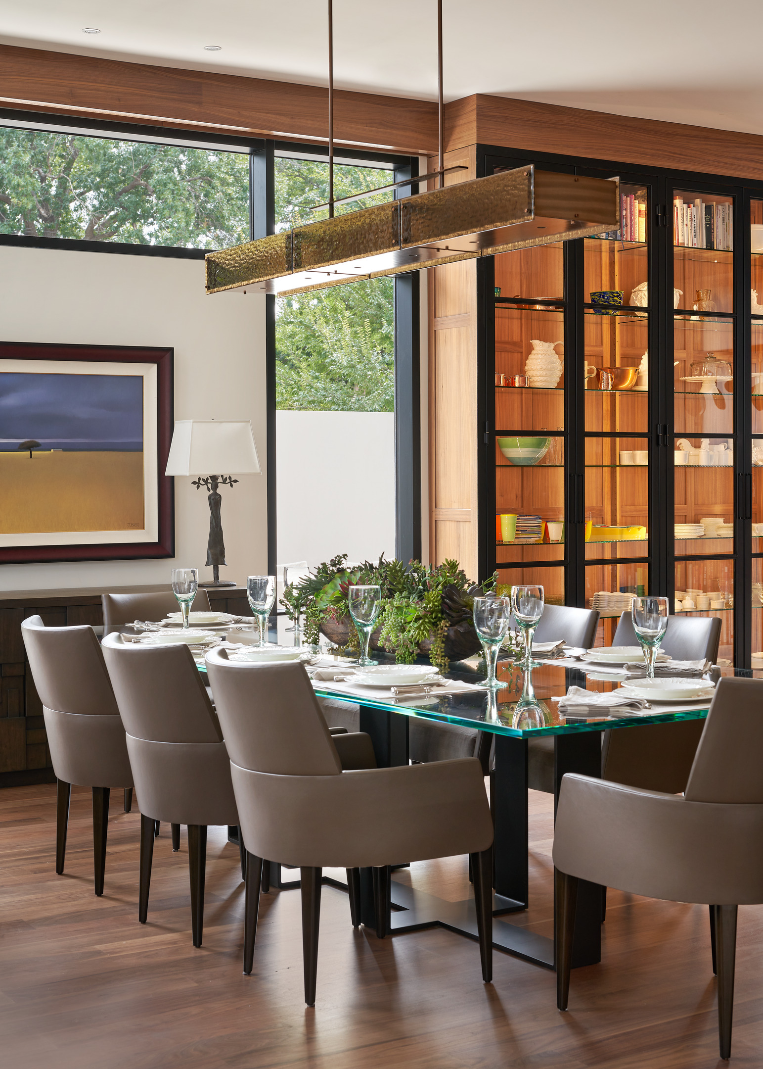 A modern dining room at the Midland Residence features a glass table, eight beige chairs, a hanging light fixture, and a bookshelf in the background. The table is set with plates, glasses, and a centerpiece.