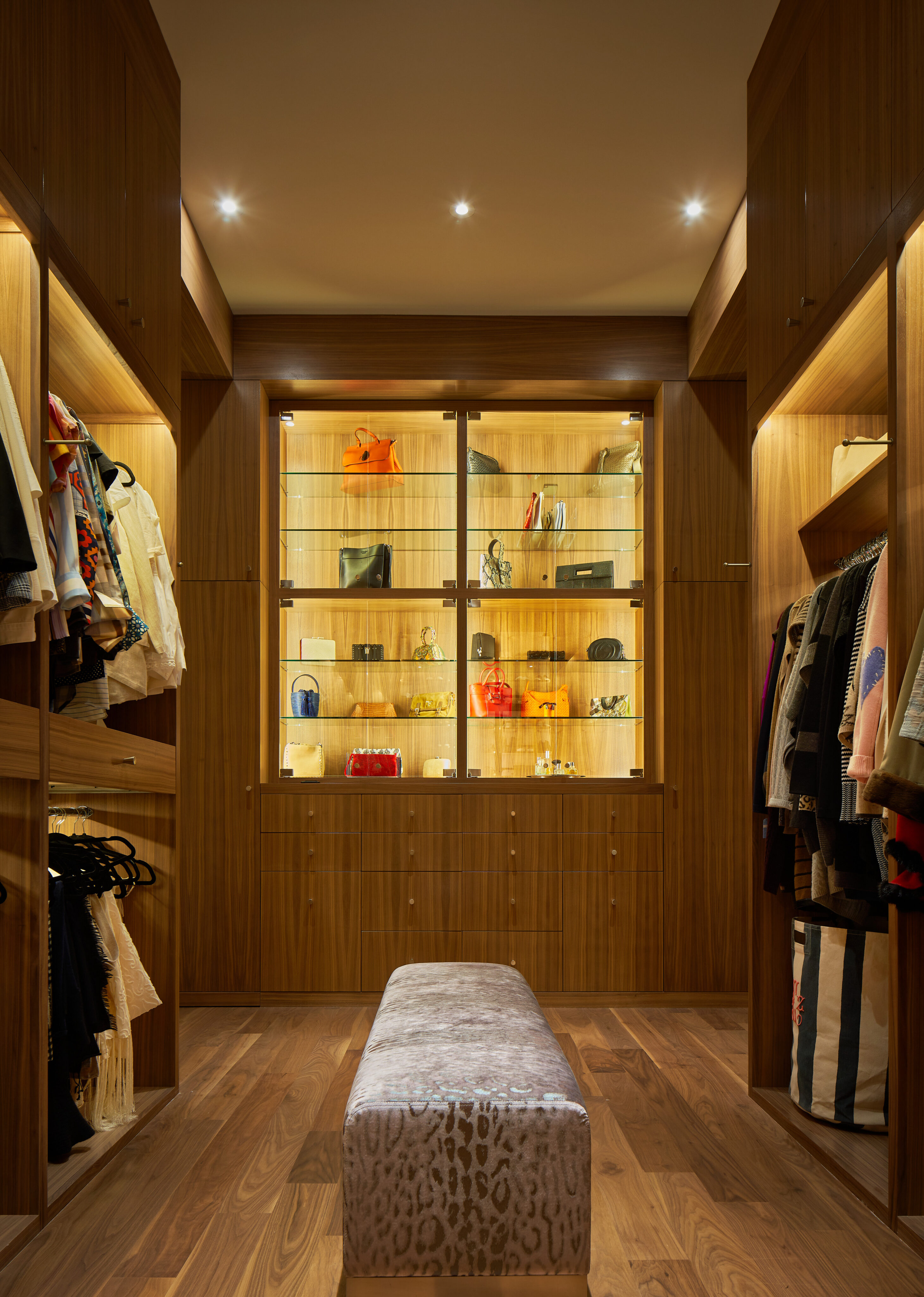 A well-lit walk-in closet in this Midland residence features wooden shelves and cabinetry, neatly organized clothes, bags, and shoes, with a bench in the center.