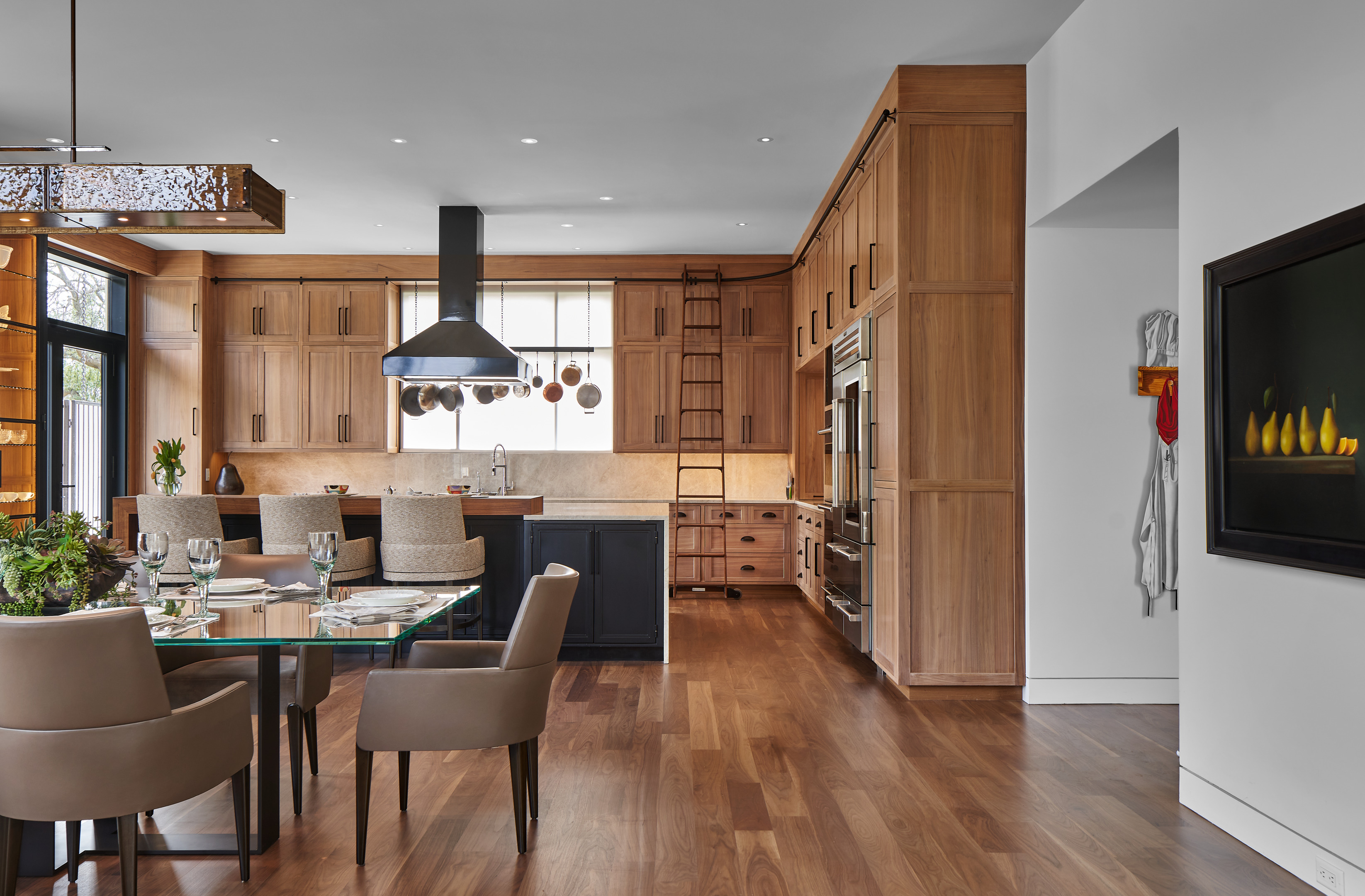 Modern kitchen and dining area at the Midland Residence features wooden cabinets, stainless steel appliances, and hardwood floors. A dining table set for four is placed near a mounted wall fireplace, creating a cozy atmosphere in this stylish Midland residence.