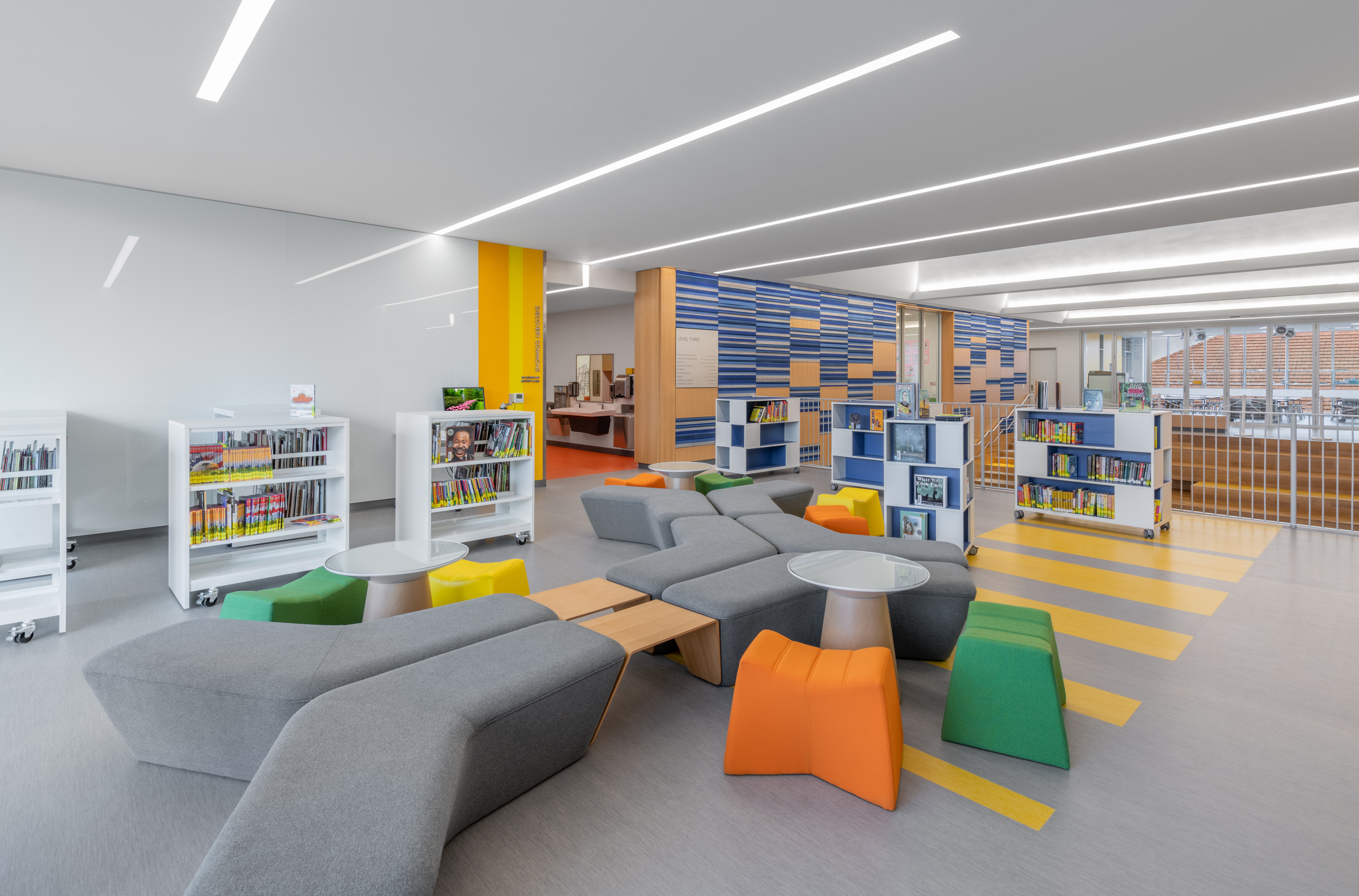 A modern library interior at ESD Lower School features colorful seating, several bookshelves filled with books, and well-lit spaces with large windows. The design includes contemporary furniture and vibrant color accents, creating an inviting atmosphere for young learners in this state-of-the-art STEM facility.