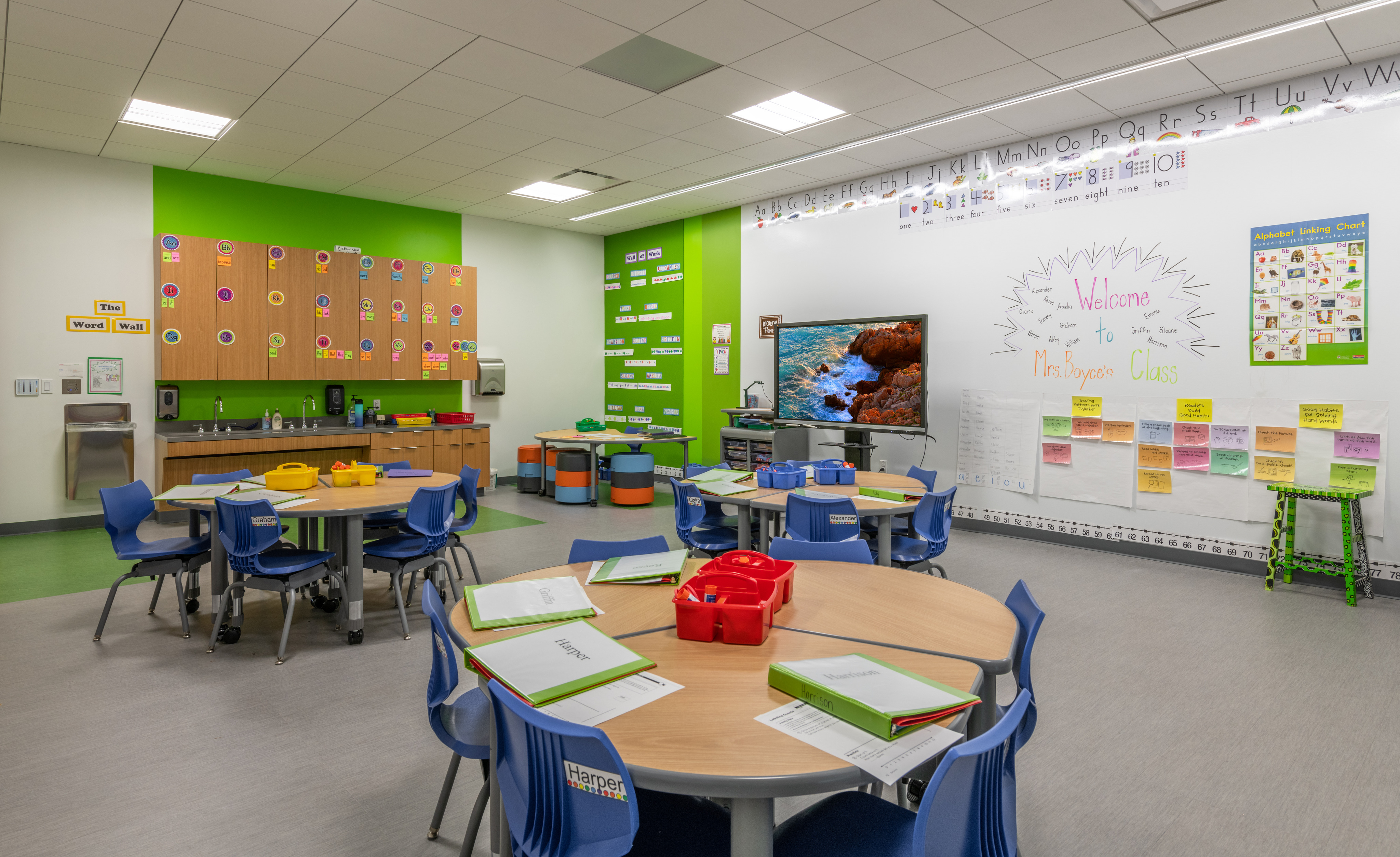 A brightly lit classroom in the ESD Lower School with round tables, blue chairs, educational posters, a whiteboard with a "Welcome" message, and various supplies neatly organized. This STEM facility fosters a vibrant learning environment for young minds.