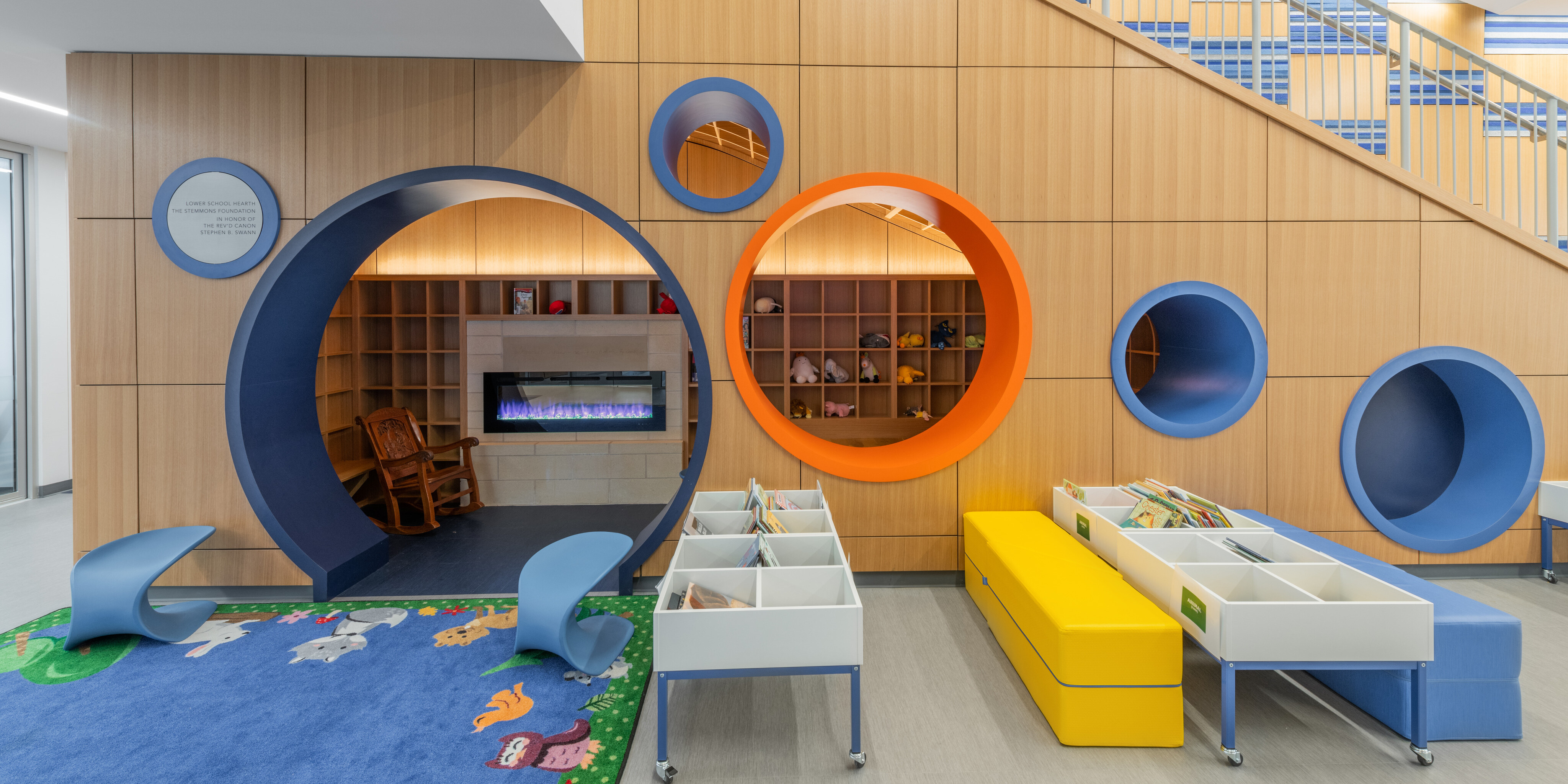 Children's reading area with colorful circular nooks, a blue rug with animals, toy bins, and a staircase in the background. The design includes wood paneling and playful, vibrant colors ideal for any Lower School STEM Facility.