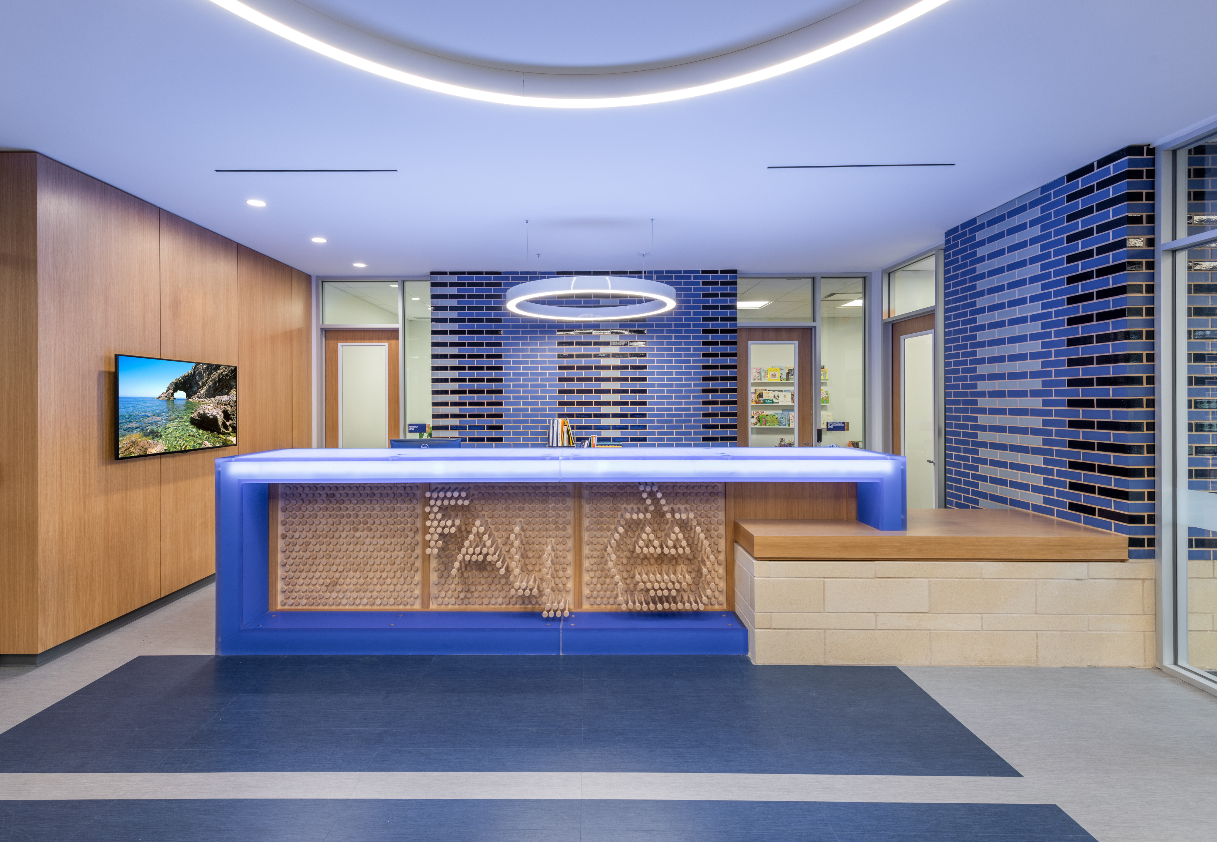 A modern reception area with a sleek design features a blue illuminated counter, a circular light fixture above, wooden accents, blue and gray tiles, and a wall-mounted screen displaying a scenic image, perfectly welcoming visitors to the cutting-edge Lower School STEM Facility.