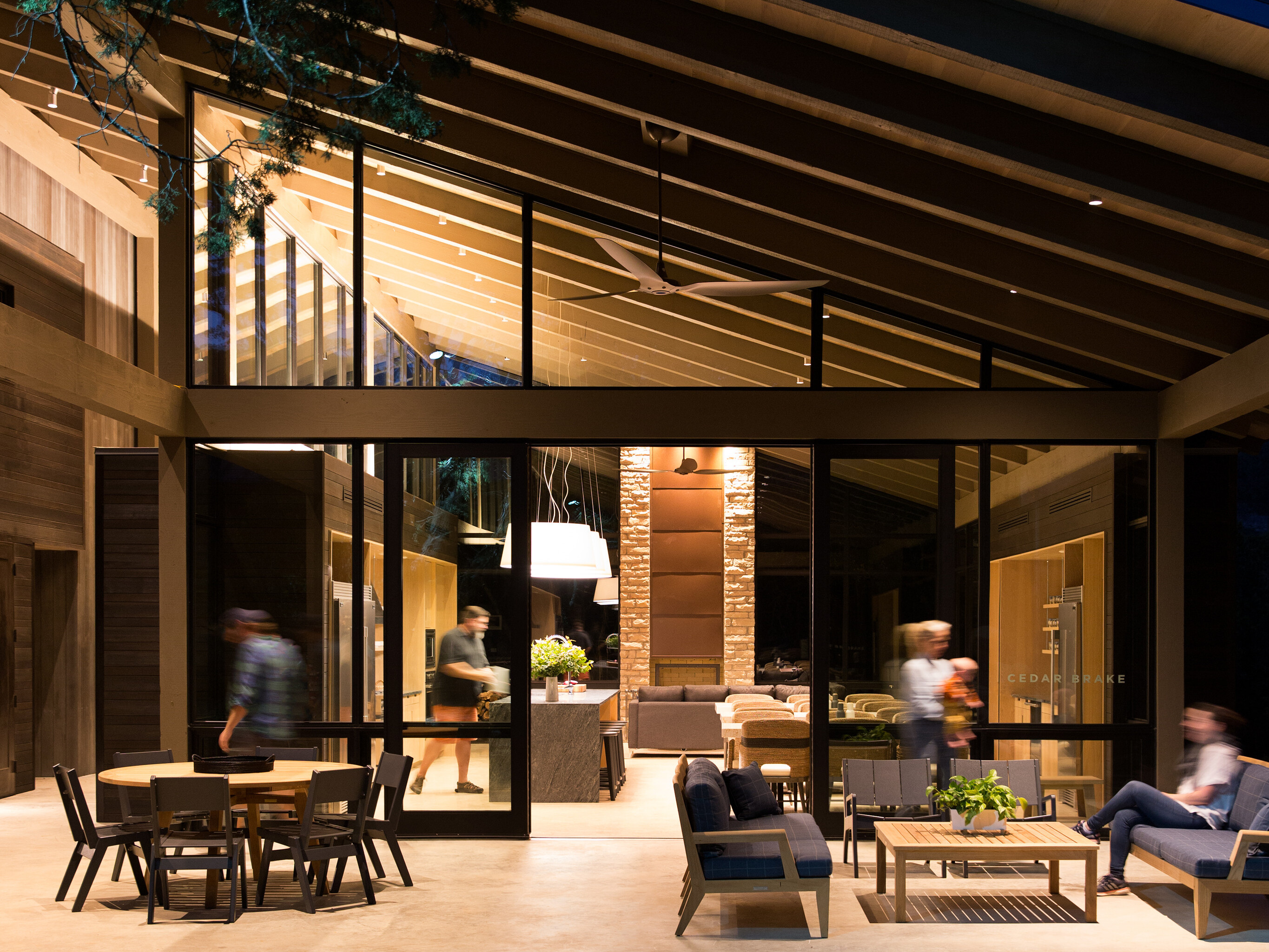 Modern house exterior at night with large glass windows showing an indoor living area. The patio, reminiscent of gatherings at Laity Lodge, features outdoor furniture and people sitting and walking around.