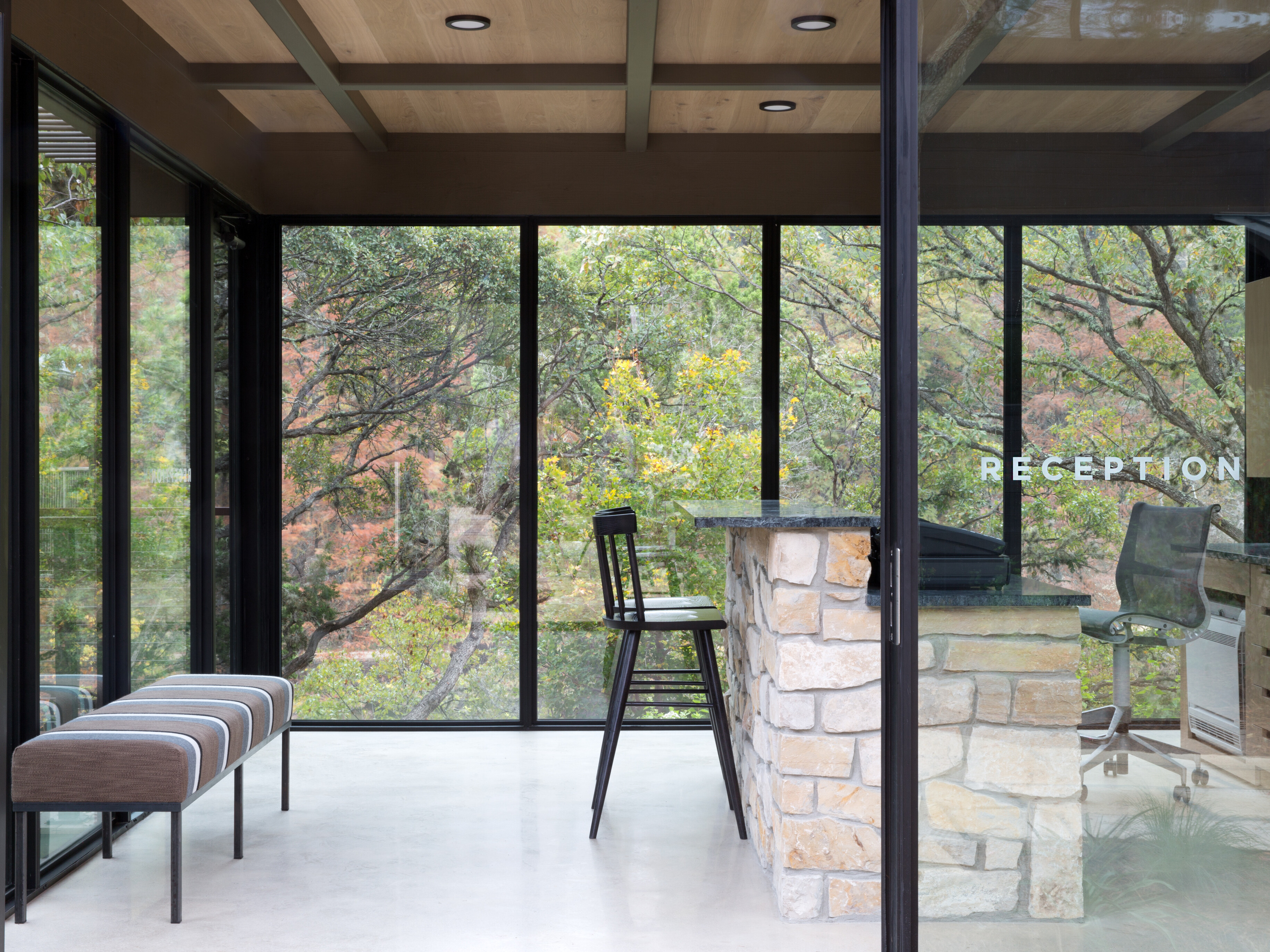 Modern reception area with glass walls, stone counter, bar stools, and waiting benches. Trees from Cedar Brake are visible outside.