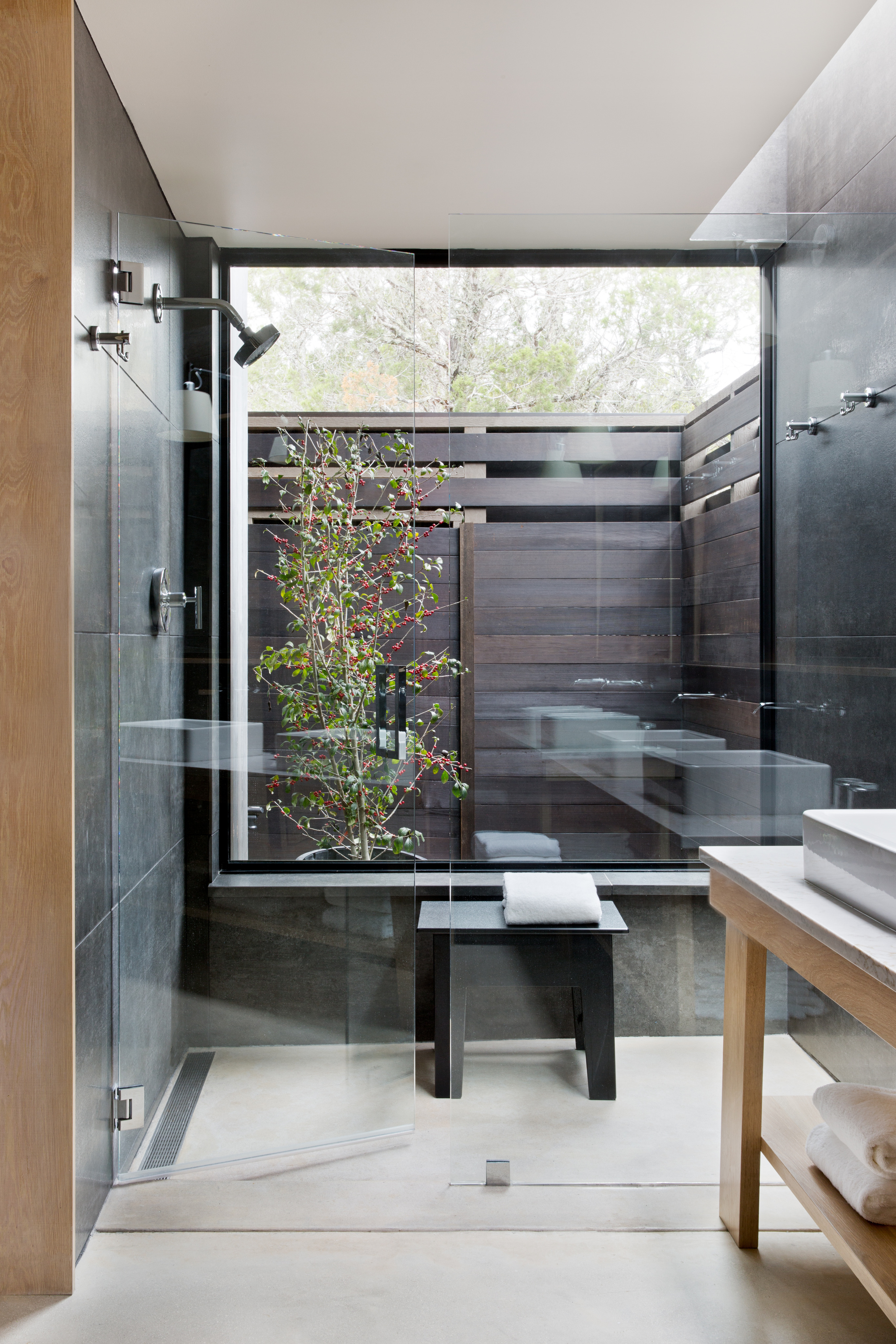 A modern bathroom features a glass-enclosed shower with a bench and a wooden privacy screen, reminiscent of Laity Lodge, with a glimpse of an outdoor plant through the partition.