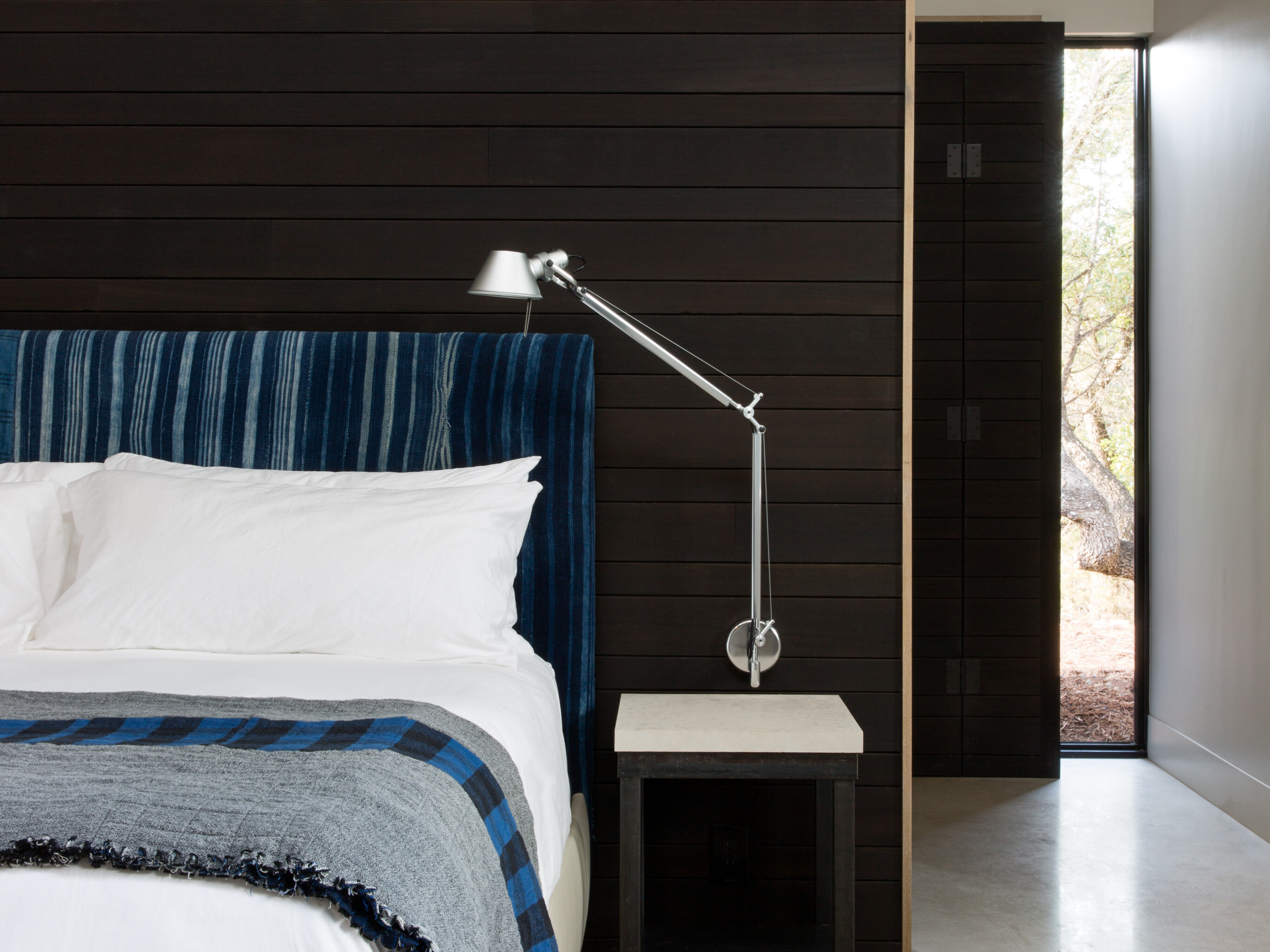 Modern bedroom with a dark wood accent wall, white bedding, and a blue padded headboard. A floor lamp, side table, and open door to the right complete the Cedar Brake minimalist decor.