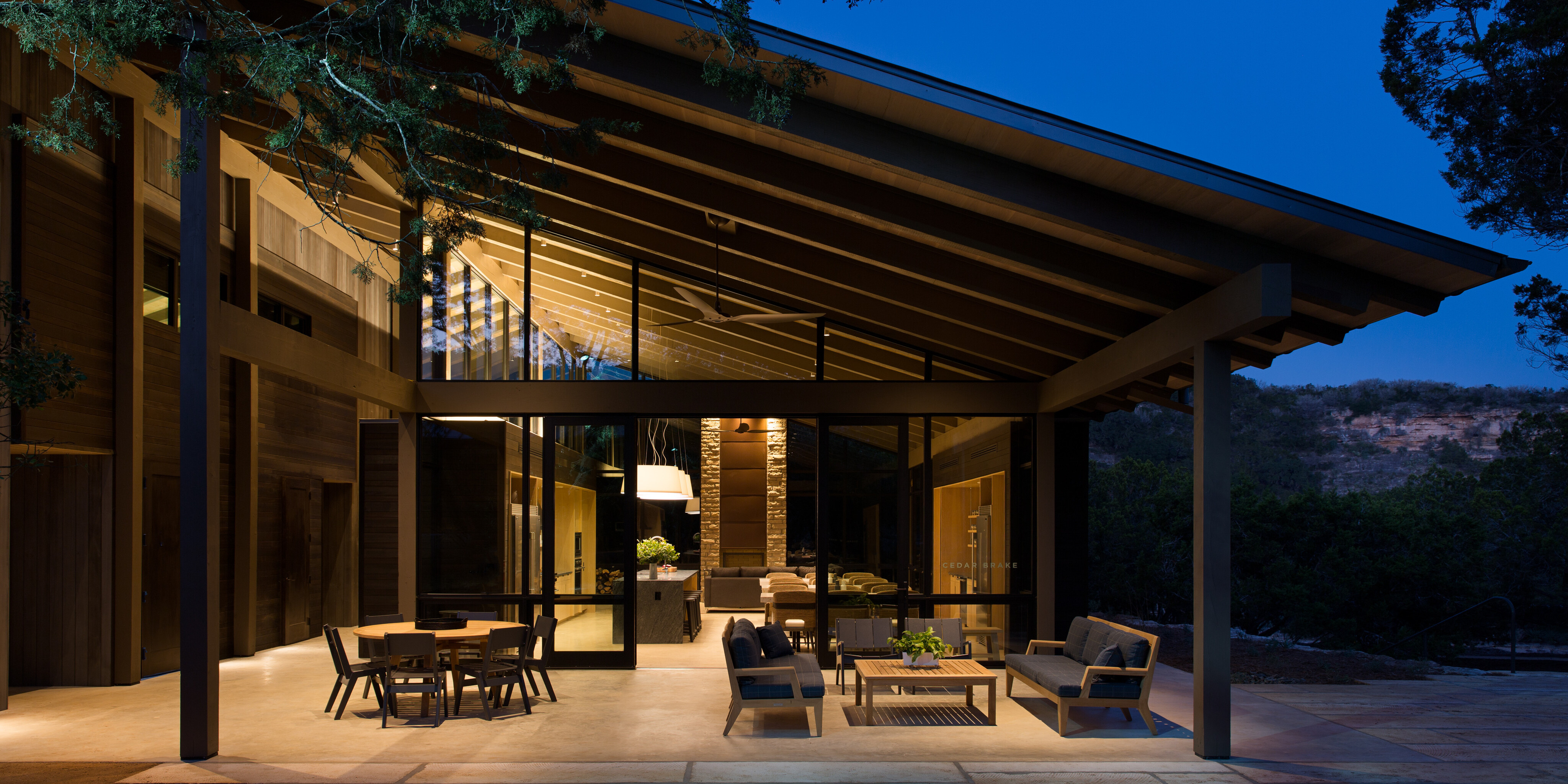 Modern house with extensive glass panels and a large overhanging roof, featuring an outdoor seating area with sofas and chairs under evening lighting. Ideal for a retreat, Cedar Brake showcases trees and hills in the background, creating a serene atmosphere reminiscent of Laity Lodge.