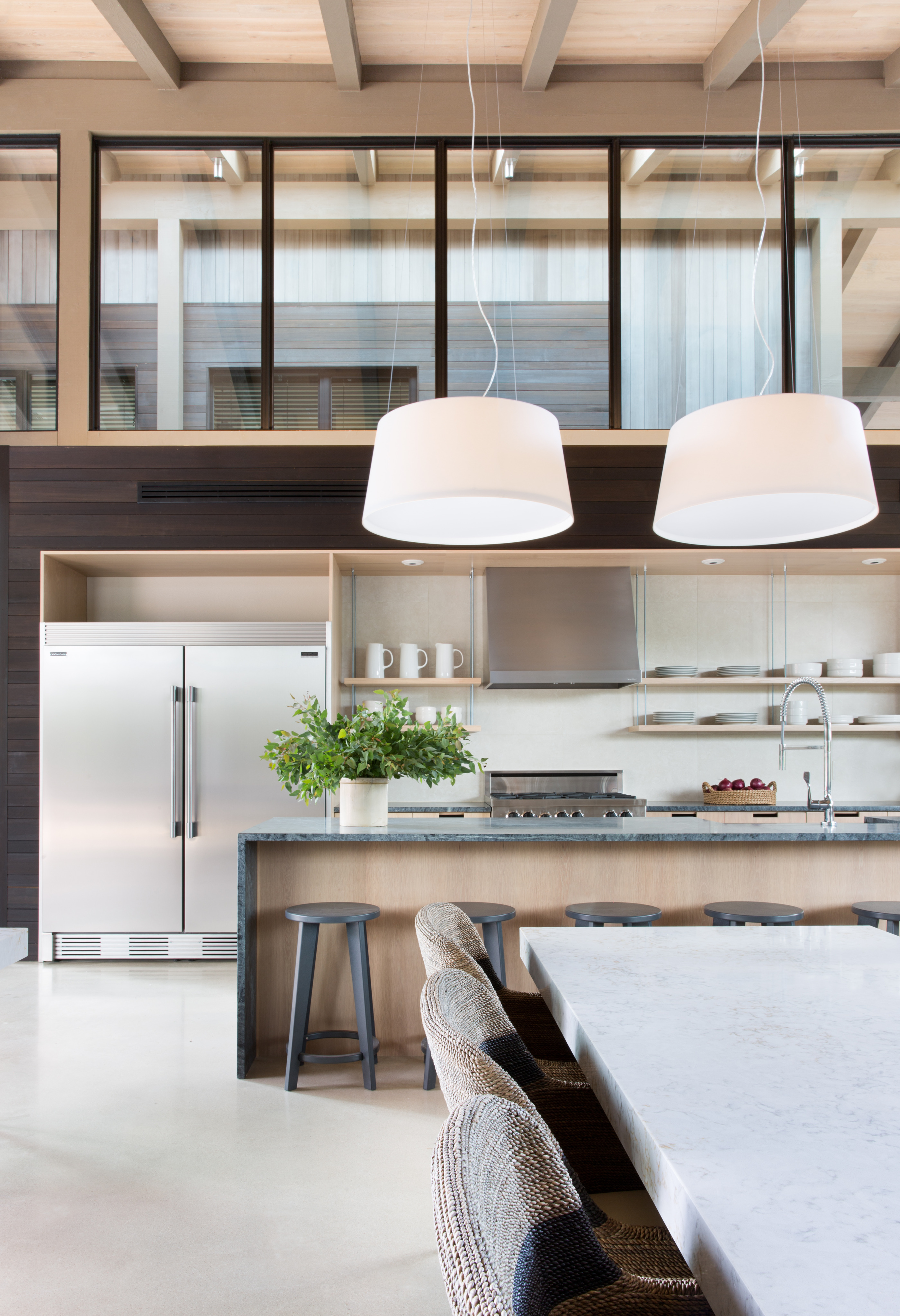 Modern kitchen with high ceilings, two hanging lights, a marble countertop island with stools, stainless steel refrigerator, gas stove, and open shelves displaying decor and kitchenware—perfect for enjoying a meal before heading out to Cedar Brake at Laity Lodge.