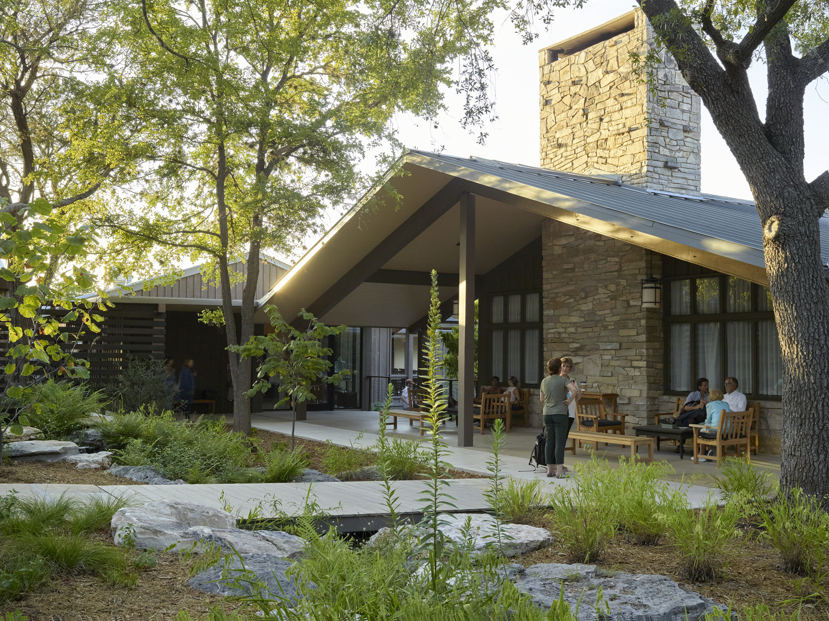 People sit on wooden chairs under the roofed entrance of a stone building surrounded by trees and greenery, nestled in the serene environment of Laity Lodge.