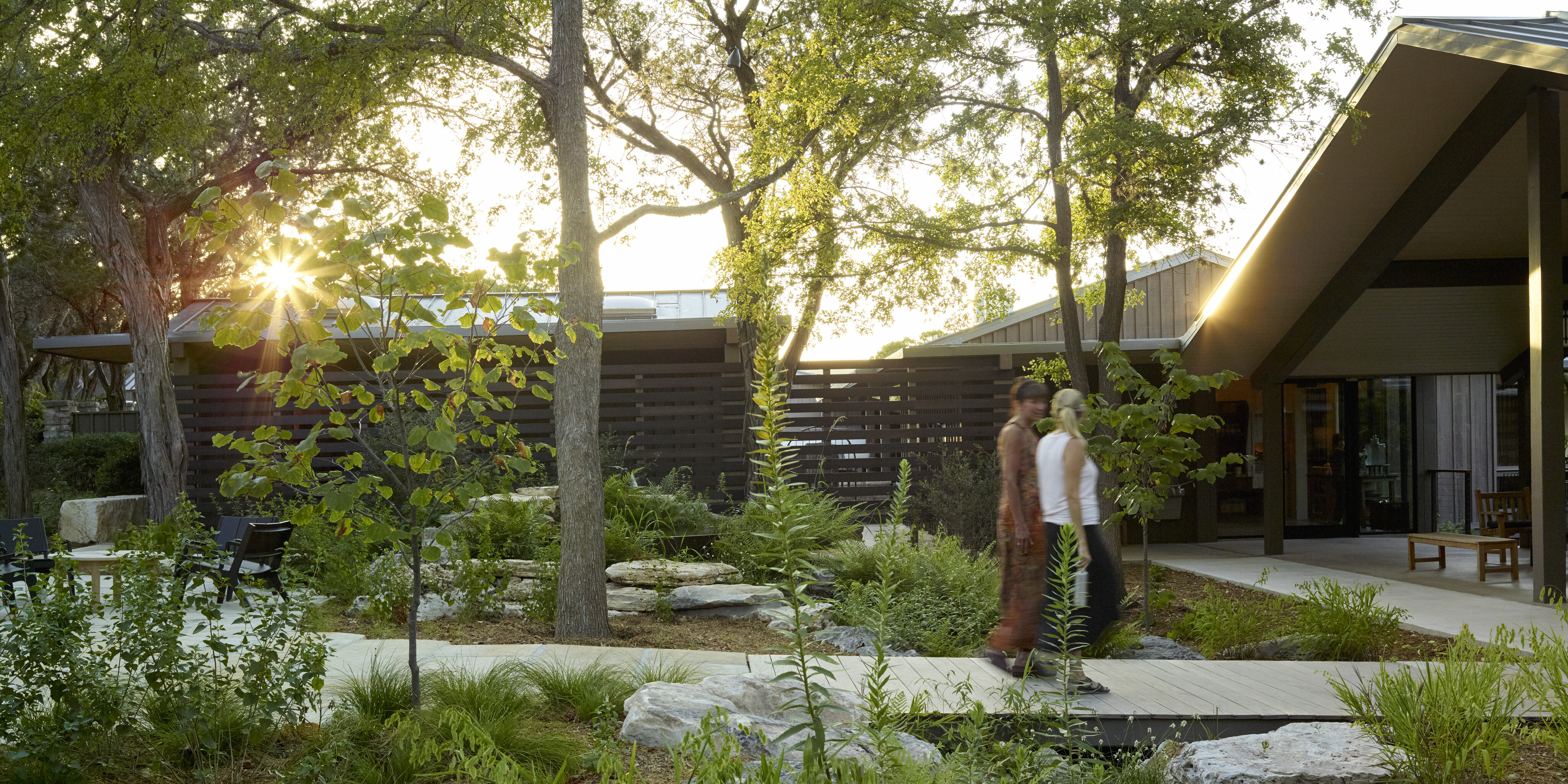 Two people walk along a path amidst lush greenery and trees near modern buildings at Laity Lodge, with the setting sun in the background.