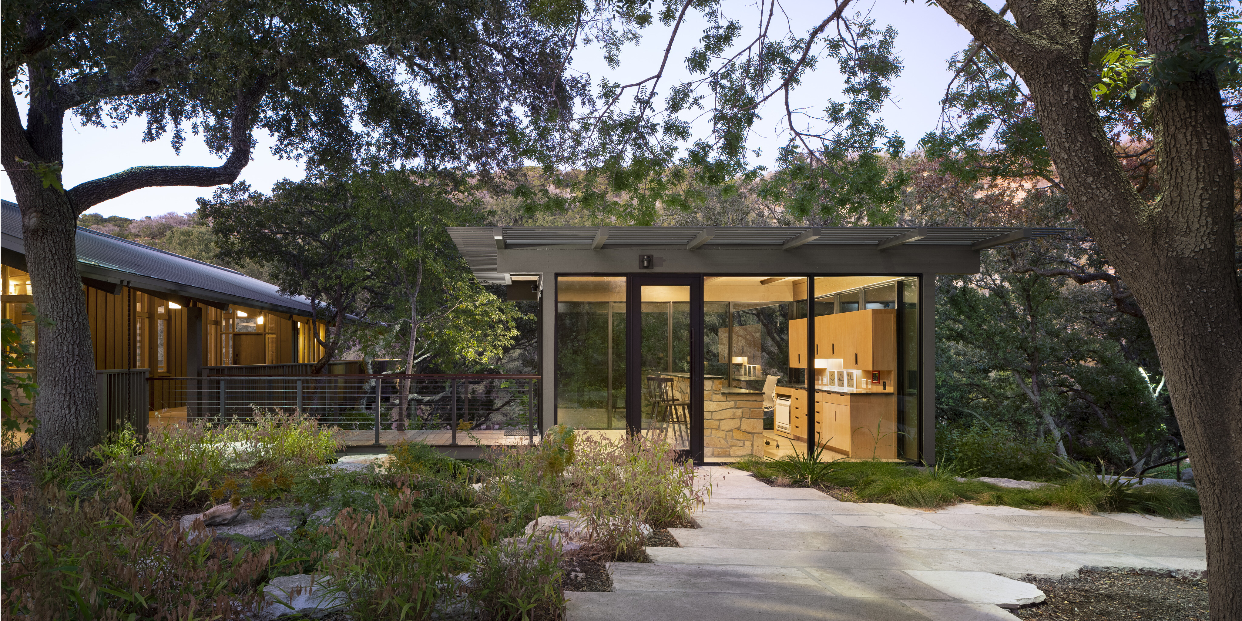 A modern glass-walled building among trees, showcasing an open kitchen inside. A pathway leads to the entrance of Cedar Brake, surrounded by natural landscaping akin to Laity Lodge's serene environment.