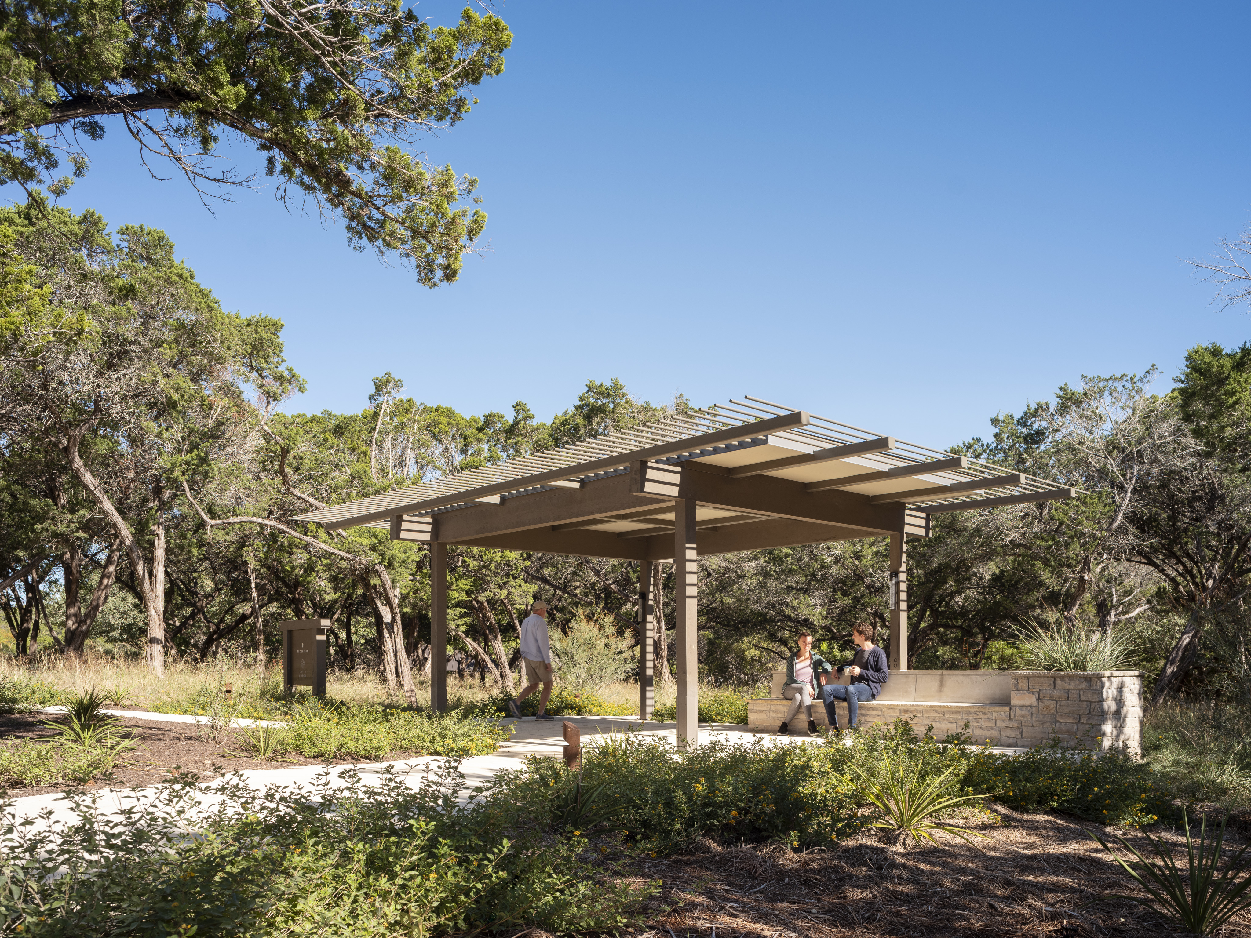 A pavilion with a slatted roof stands in the Cedar Brake area of Laity Lodge, surrounded by greenery and trees. Two people sit on a stone bench under the pavilion while one person walks nearby. The sky is clear and blue.