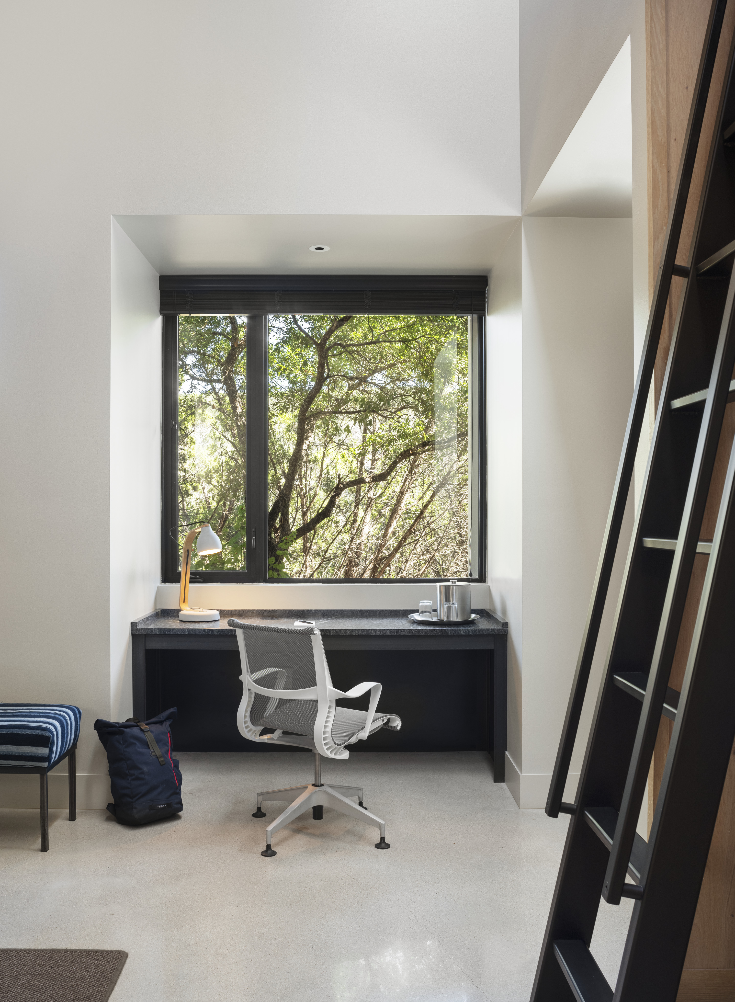 Modern home office space with a desk and chair positioned in front of a large window offering a view of Cedar Brake's serene trees. A lamp and a few objects adorn the desk, while a staircase is visible on the right.